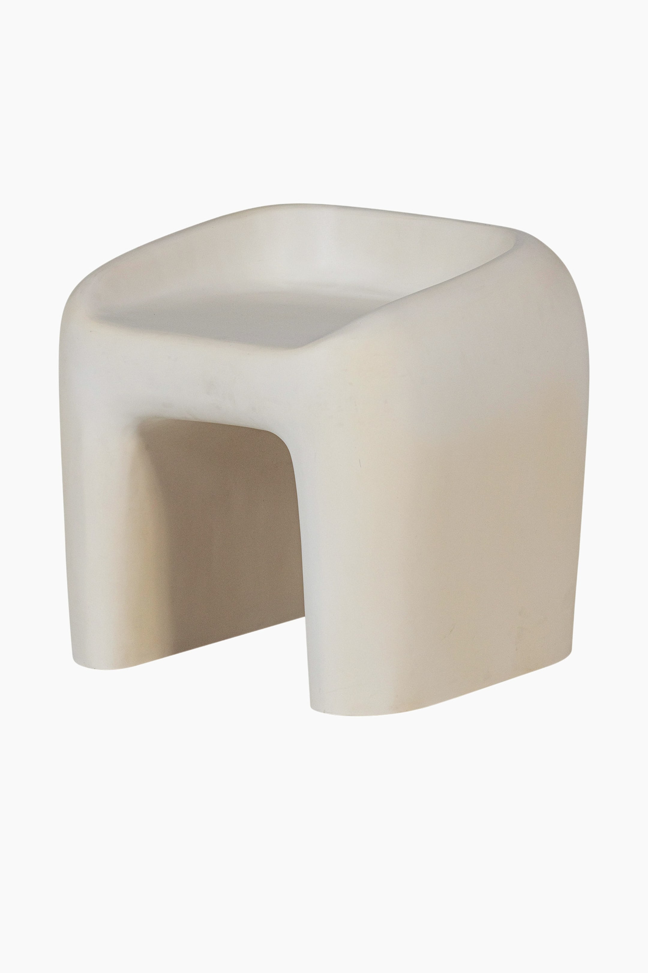 Zobacz większy obraz: Jasny beżowy, monolityczny stołek prezentuje swoją przednio-lewą stronę, charakteryzującą się podstawą w kształcie litery U, która wspiera delikatnie wklęsłe siedzisko z zaokrąglonymi krawędziami.