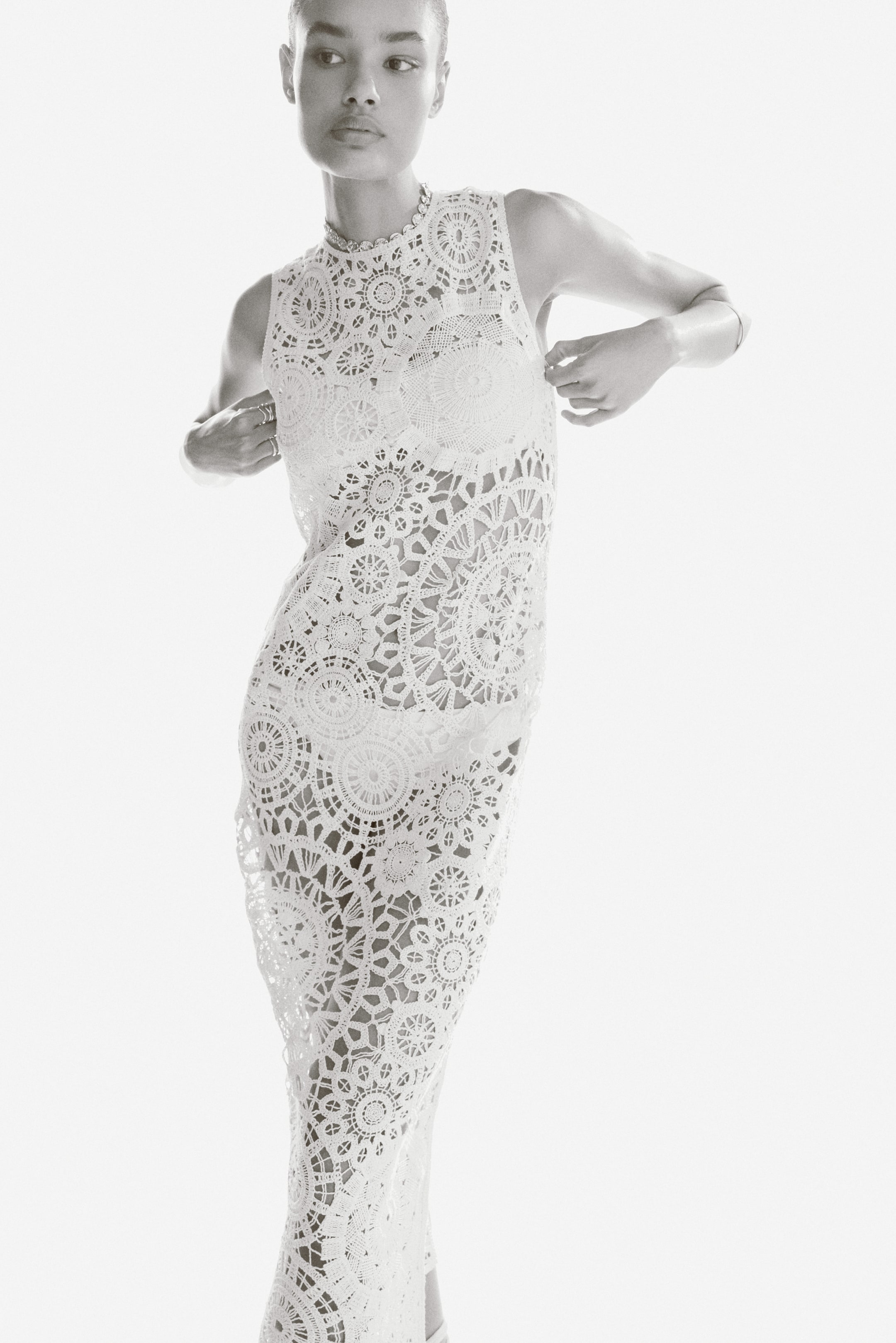 View larger image: The model is wearing a white, long, sleeveless dress crafted in a crochet-look knit. This dress showcases a round neckline, a slim fit, detailed openwork patterns, and a scalloped hem, offering a subtle see-through effect. The model is gently adjusting the sides of the dress with both hands.