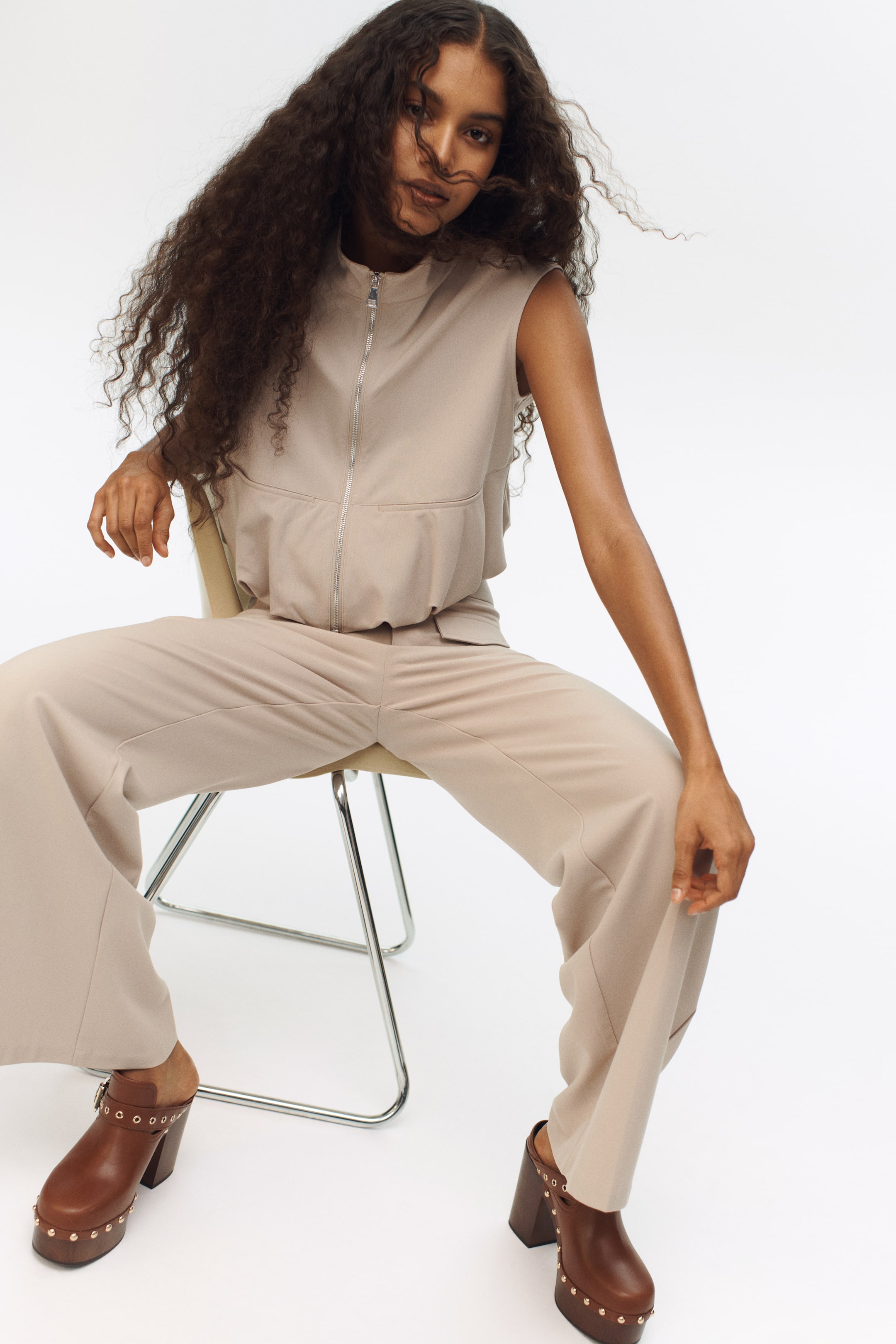 Zobrazit větší obrázek: Béžový top bez rukávů, s vysokým límcem a celoplošným stříbrným zipem, je kombinován s odpovídajícími kalhotami s širokými nohavicemi, které nabízejí uvolněný střih. Hnědé dřeváky s robustním podpatkem, platformovou podrážkou a zlatými cvočky a sponou na pásku dokončují tento outfit.