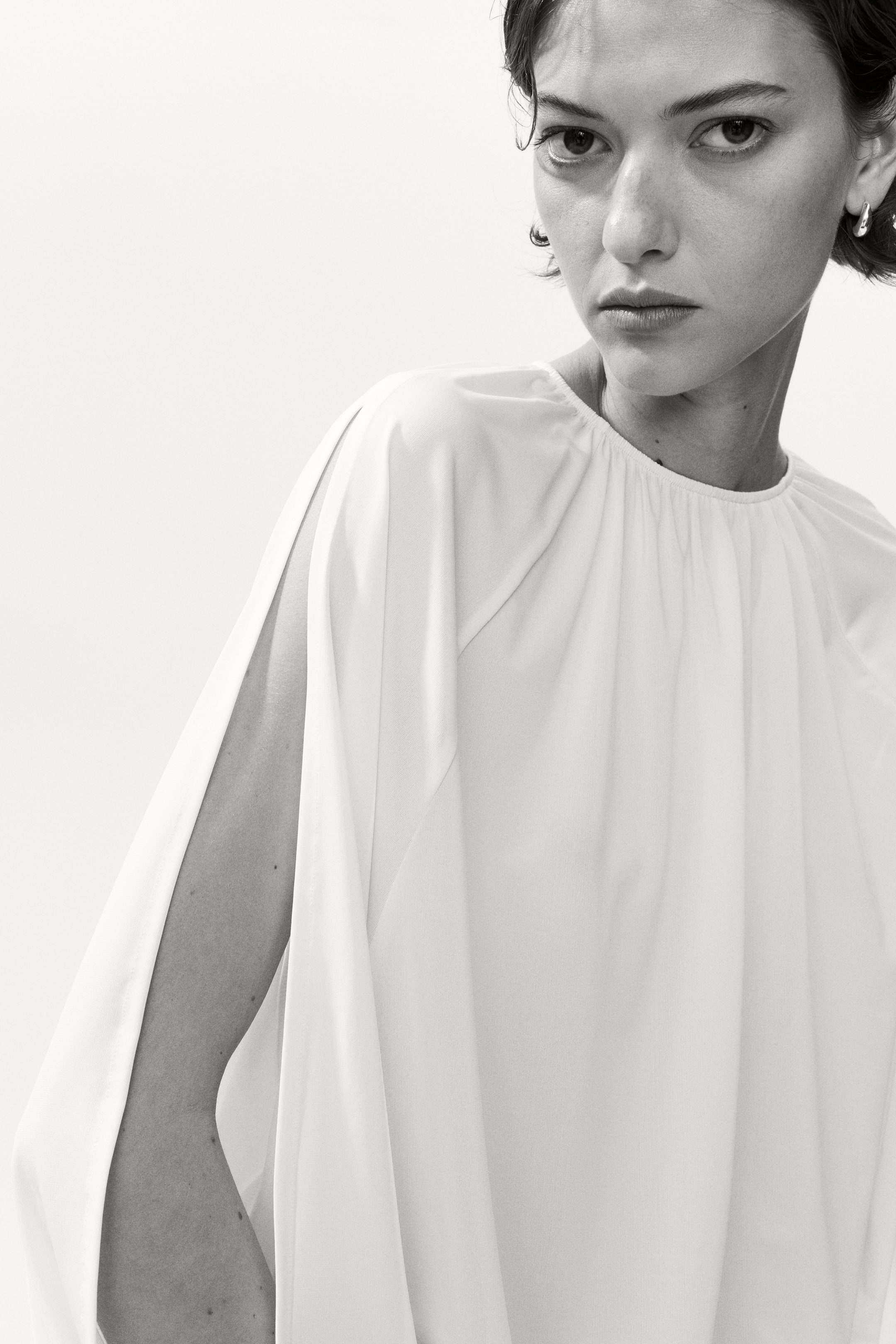 View larger image: The model is wearing a loose-fit top, showcasing a round neckline with distinct gathers and long raglan sleeves that have an open outer side, revealing her arm. The top presents a voluminous silhouette.