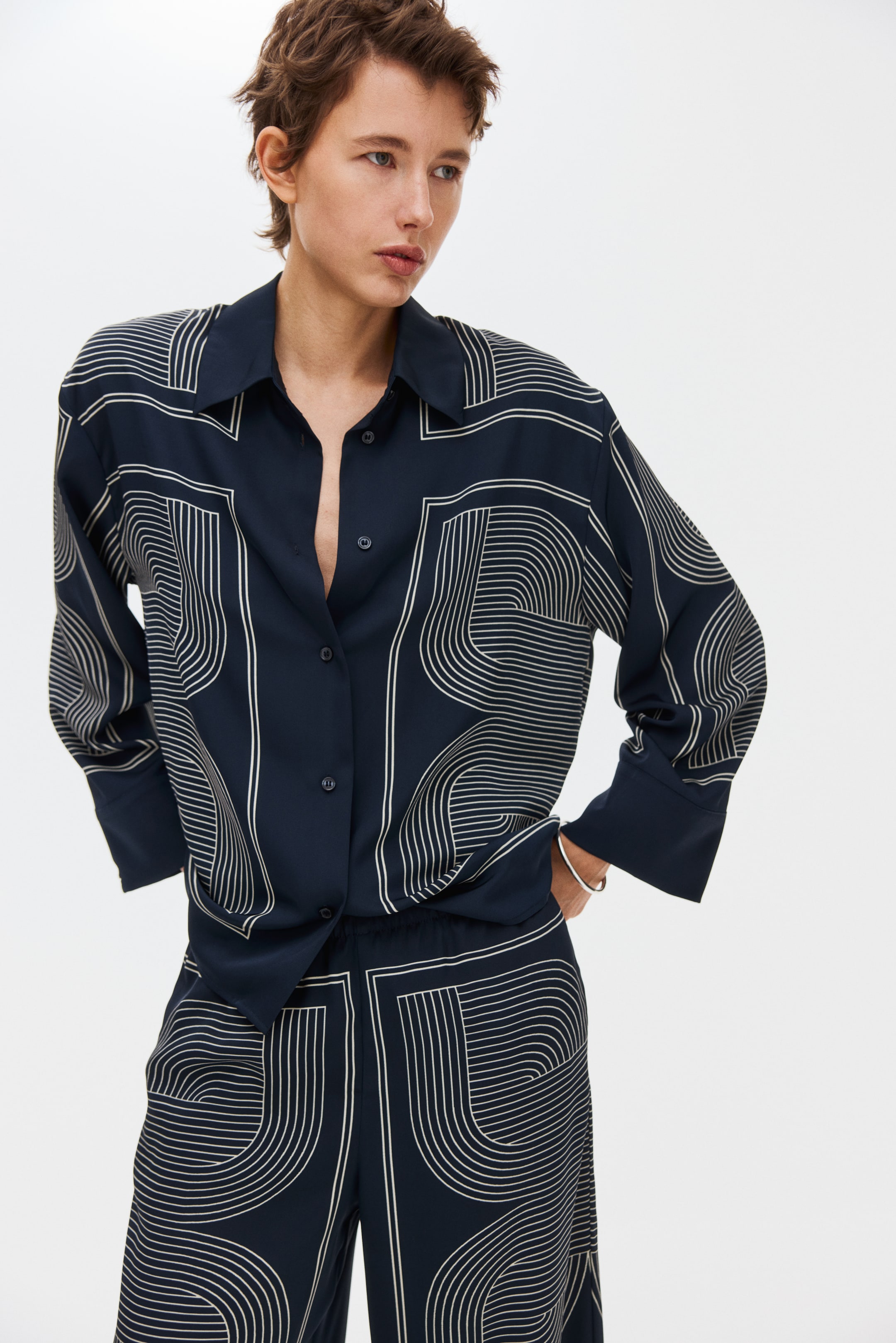 Größeres Bild ansehen: Von vorne präsentiert zeigen ein marineblaues Hemd mit Kragen und eine dazu passende Hose mit weitem Bein ein sich wiederholendes, helles, geometrisches Muster aus parallelen Linien, die Bögen und Winkel bilden. Das Hemd zeigt lange Ärmel mit breiten Manschetten und vorne eine teilweise aufgeknöpfte Knopfleiste, während die Hose mit einem elastischen Bund versehen ist.