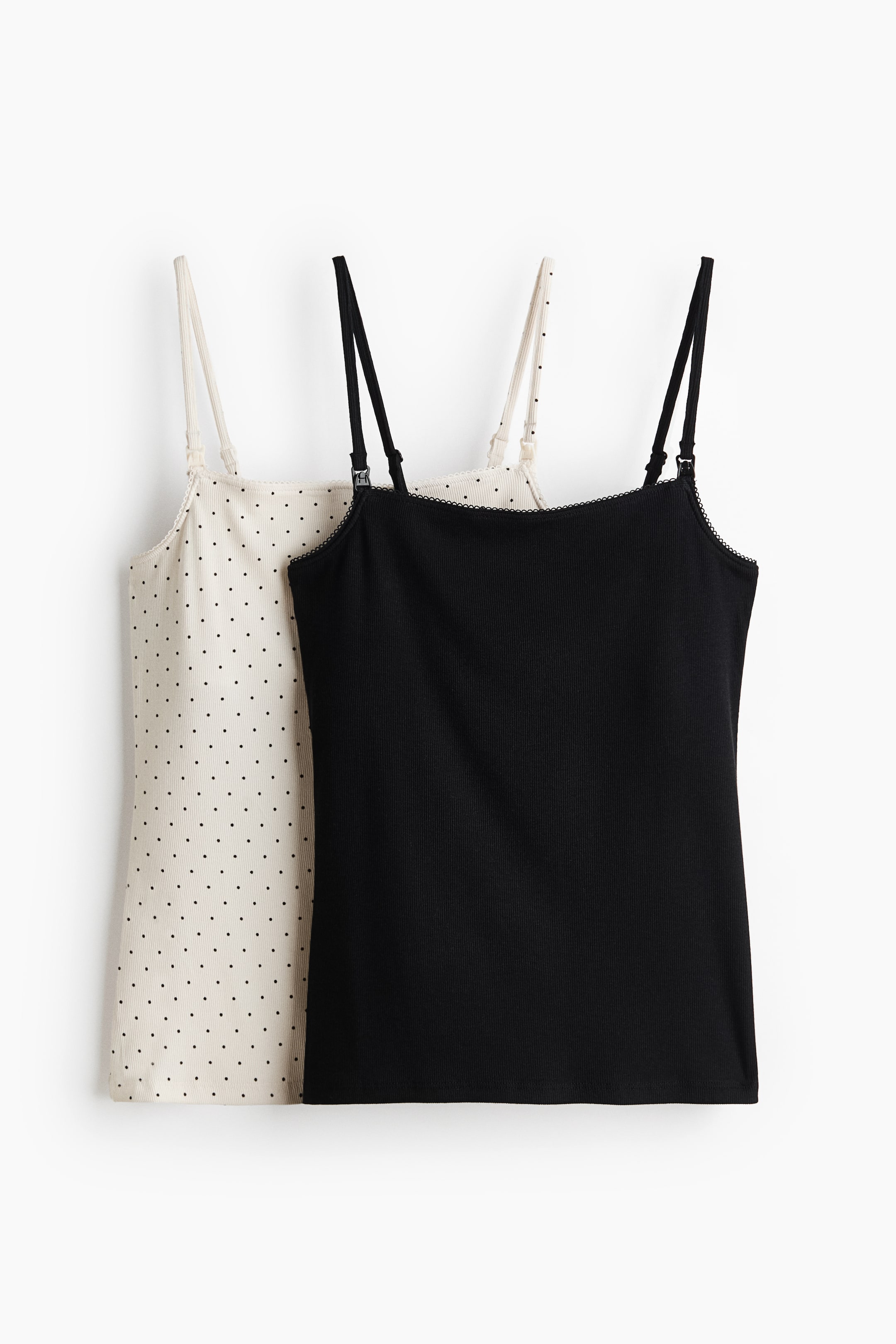Größeres Bild ansehen: Zwei gerippte Jersey-Stilltops werden aus einer leicht schrägen Frontansicht präsentiert. Ein schwarzes Top mit einem geraden, mit Picot-Besatz versehenen Ausschnitt und schmalen, verstellbaren Trägern mit funktionalen Verschlüssen ist teilweise vor einem cremeweißen Top mit schwarzen Punkten positioniert.