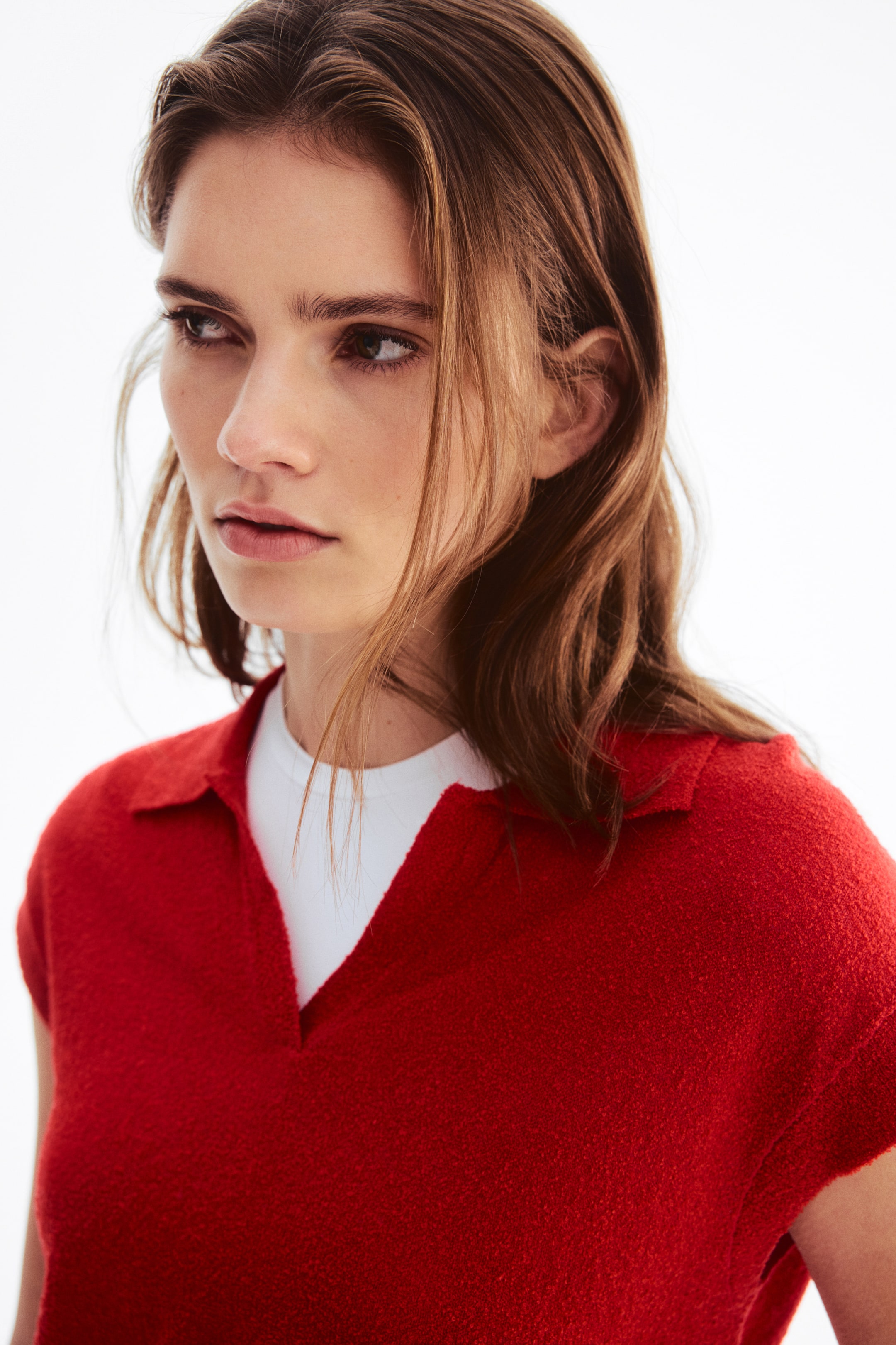 Ver imagem maior: Um top vermelho de malha texturizada de mangas curtas, com gola aberta e decote em V, está a ser usado sobre uma t-shirt branca e é apresentado num ângulo ligeiramente de lado.