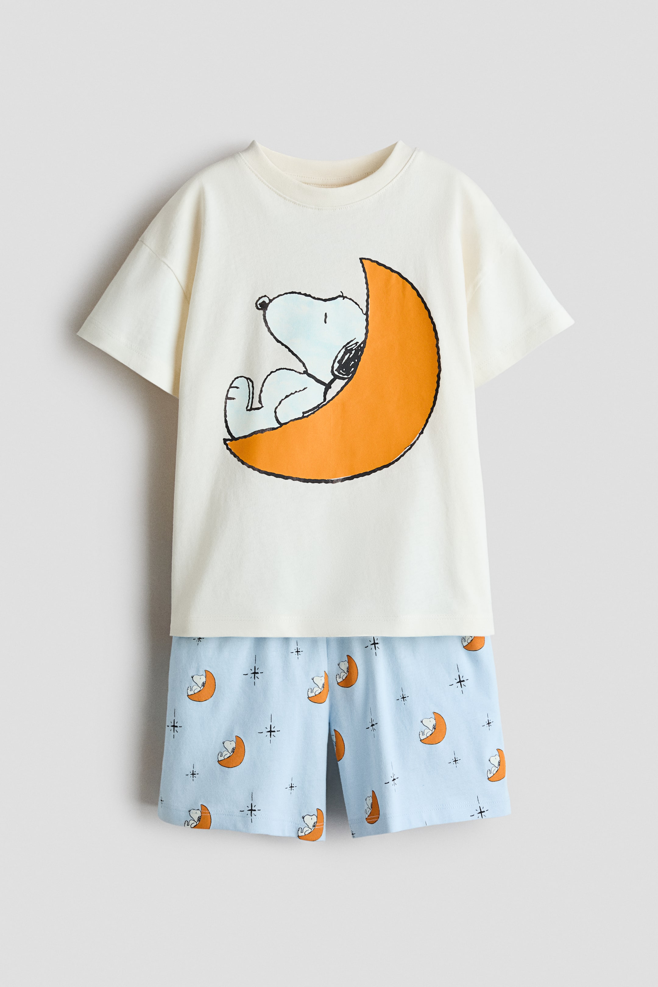 View larger image: A cream-coloured short-sleeved t-shirt and light blue shorts pyjama set is presented front-facing. The t-shirt showcases a large graphic of Snoopy sleeping on an orange crescent moon, while the shorts display a repeating pattern of small Snoopy-on-moon motifs and tiny cross-shaped stars.
