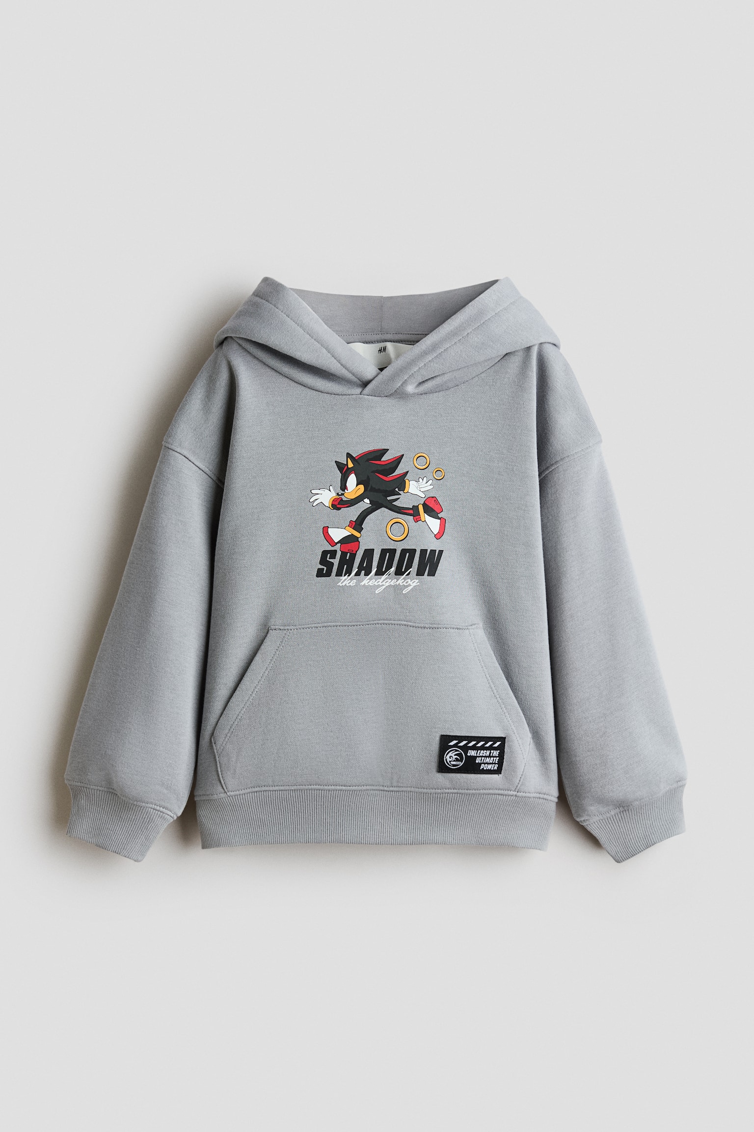 Een lichtgrijze hoodie met losse pasvorm, frontaal weergegeven, met op de borst een opdruk van Shadow the Hedgehog en de tekst "SHADOW the hedgehog". Het kledingstuk heeft verlaagde schouders, een capuchon met overslag aan de voorkant, een kangoeroezak met een klein zwart patchje en geribde boorden en zoom.