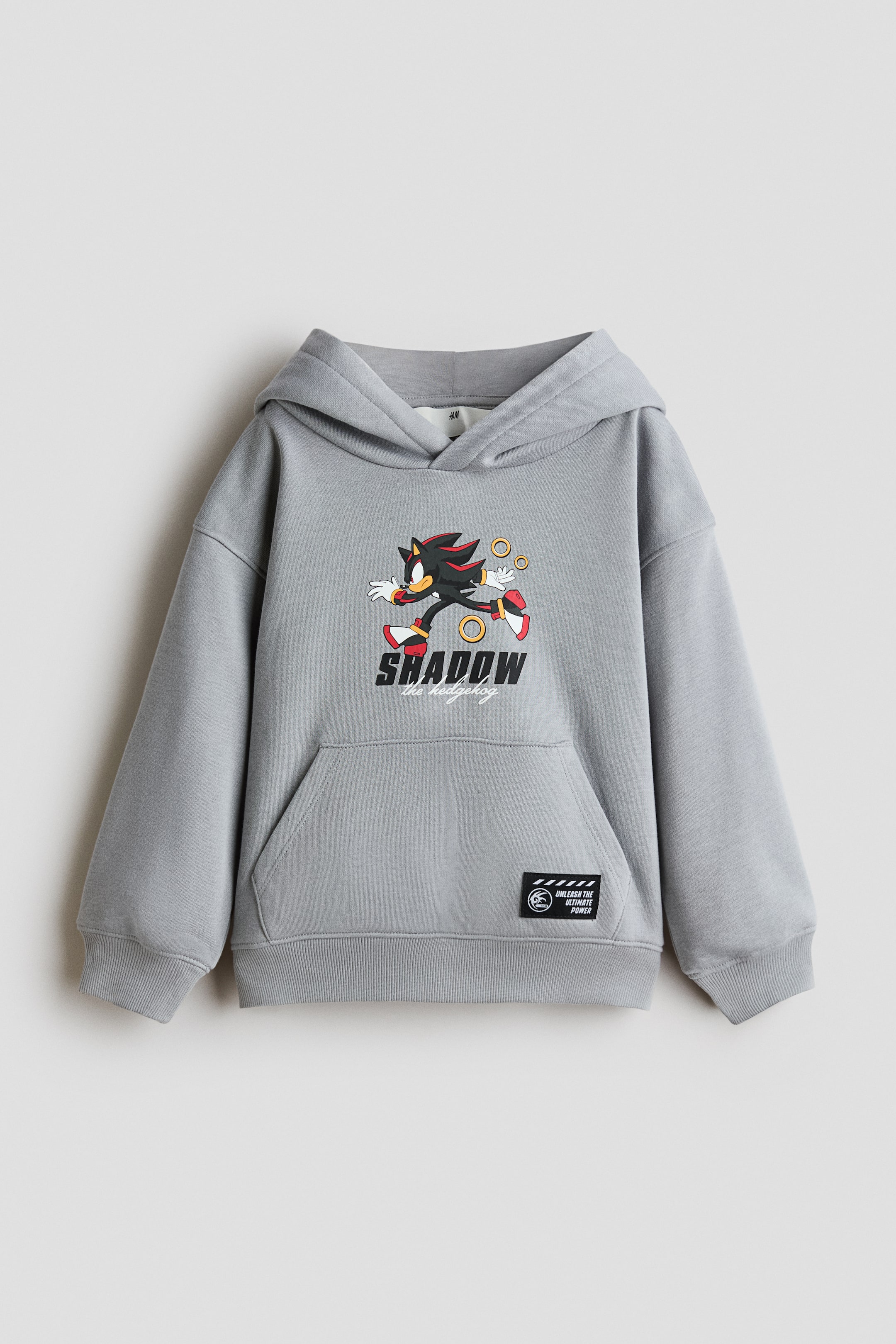 View larger image: A light grey loose-fit hoodie is shown from the front, showcasing a printed graphic of Shadow the Hedgehog and the text "SHADOW the hedgehog" on the chest. The garment features dropped shoulders, a wrap-front hood, a kangaroo pocket with a small black patch, and ribbed cuffs and hem.