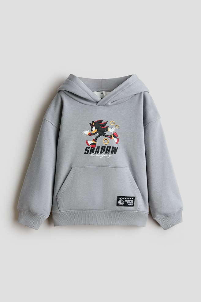 Una sudadera holgada de color gris claro está de frente, mostrando en el pecho un gráfico impreso de Shadow the Hedgehog y el texto "SHADOW the hedgehog". La prenda presenta hombros caÃdos, capucha cruzada al frente, un bolsillo tipo canguro con un pequeño parche negro y puños y dobladillo acanalados. - 1