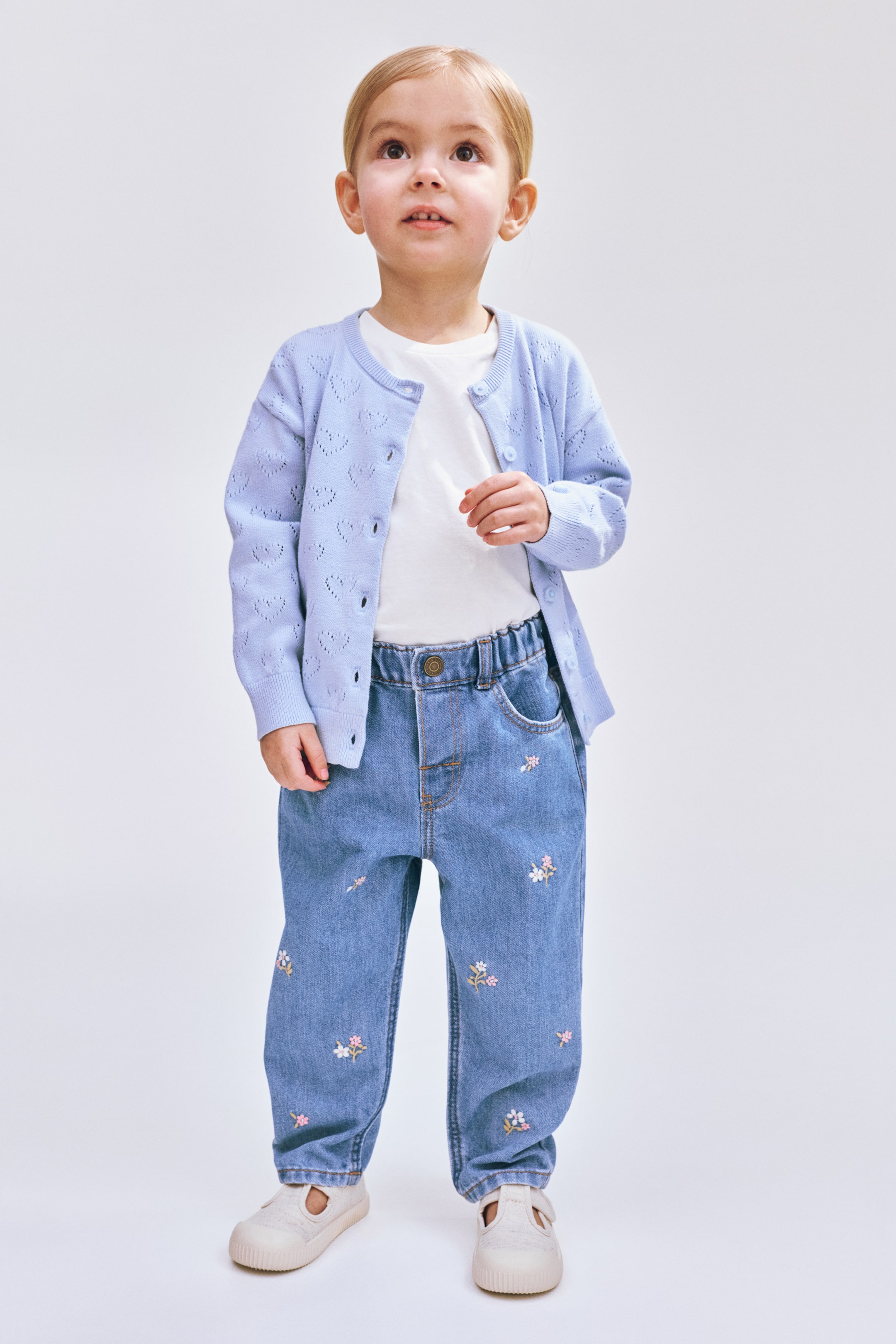 View larger image: A light blue openwork heart-patterned cardigan, featuring a round neck, ribbed cuffs, and front button closure, is worn over a white T-shirt. Light blue denim jeans with an elastic waist, front pockets, and scattered floral embroidery are also worn.