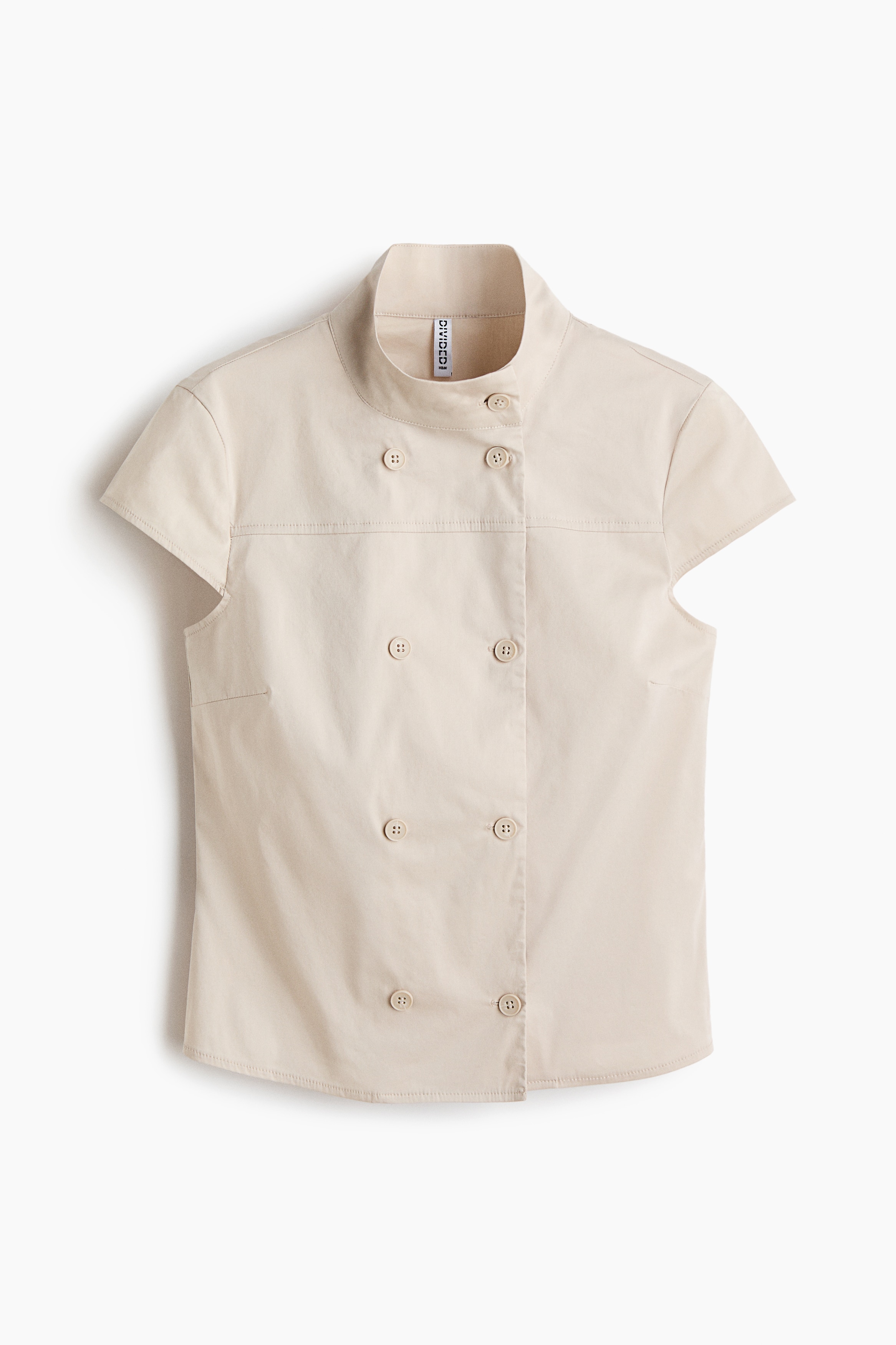 Un haut beige ajustÃ© avec un col montant, une patte de boutonnage Ã  deux rangÃ©es, un empiÃ¨cement devant et des manches courtes est portÃ© par le mannequin. Le haut est associÃ© Ã  un jean en denim bleu, le mannequin ayant les mains dans les poches.
