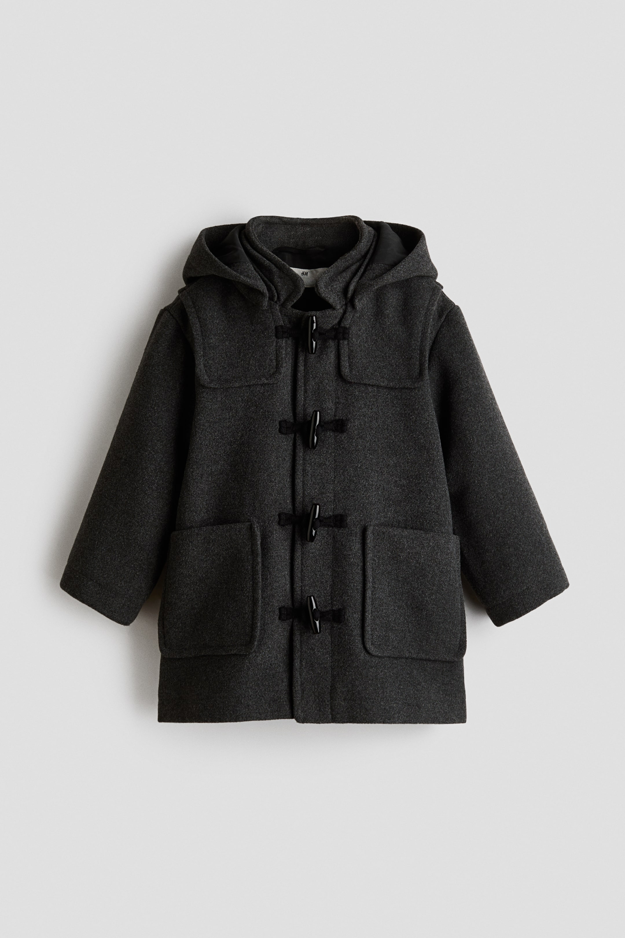 Ver imagen más grande: Se muestra de frente un duffle coat de color gris oscuro en tejido afieltrado, con capucha forrada, canesúes en los hombros y cuatro cierres tipo toggle negros en el centro. Dos grandes bolsillos de parche abiertos se sitúan en la parte delantera inferior.