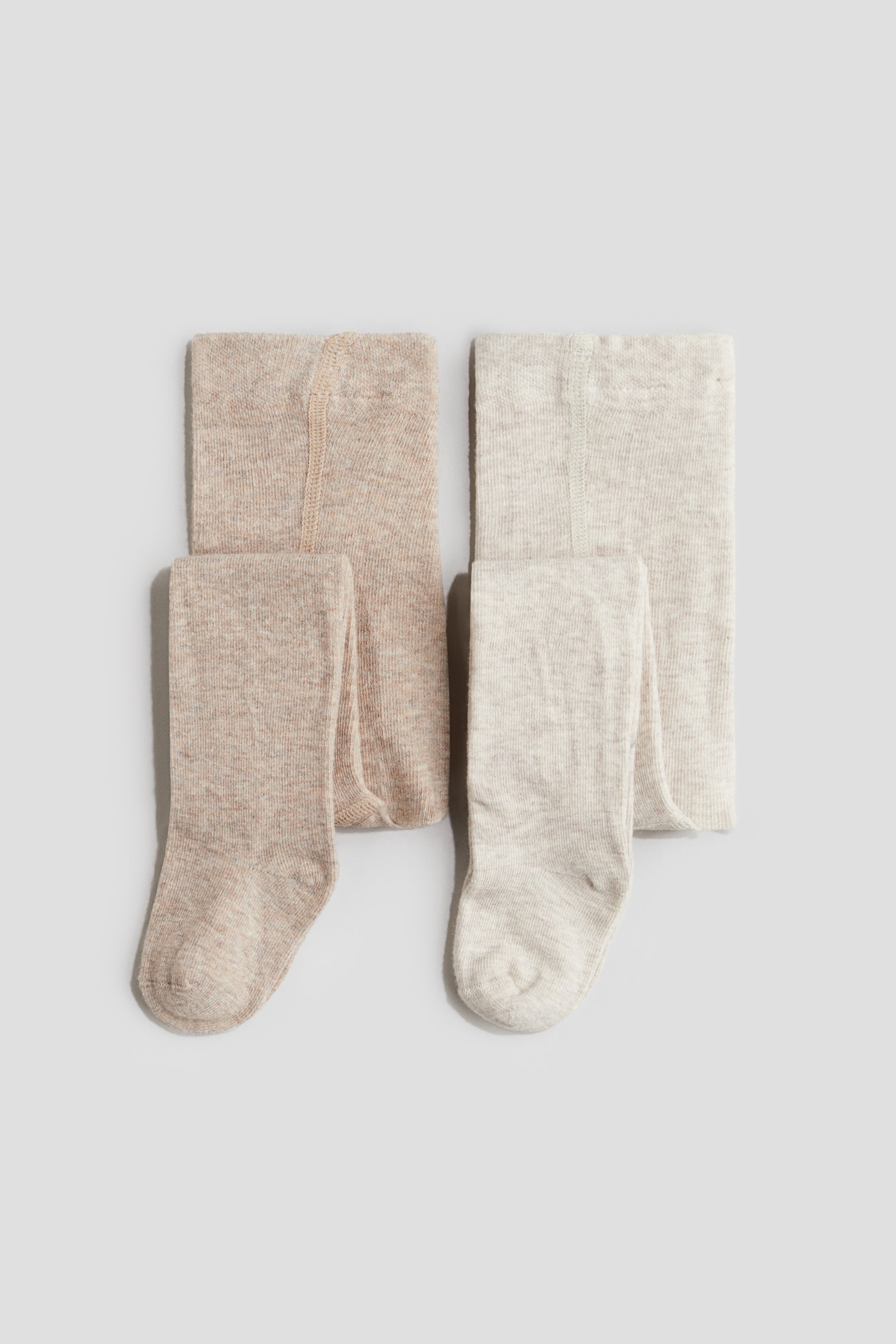 Ampliar la imagen: Dos pares de mallas en una suave mezcla de algodón de tejido fino están colocados en posición horizontal, uno junto al otro, con un par en beige jaspeado y el otro en crema jaspeado. Ambos pares están doblados para mostrar la cintura elástica en la parte superior, con una pierna de cada par colgando hacia abajo para mostrar la construcción del pie.