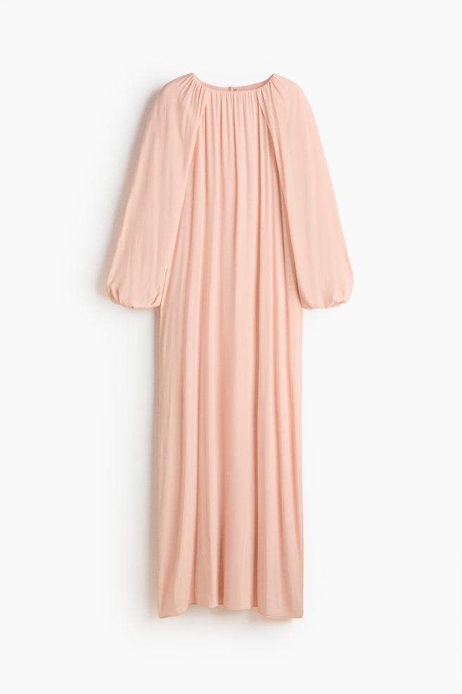 A light peach dress is displaying its front, featuring a gathered round neckline and long, voluminous sleeves that are cinched at the cuffs. The fabric is flowing loosely from the neckline, forming soft vertical pleats down the length of the maxi-style garment. - 1