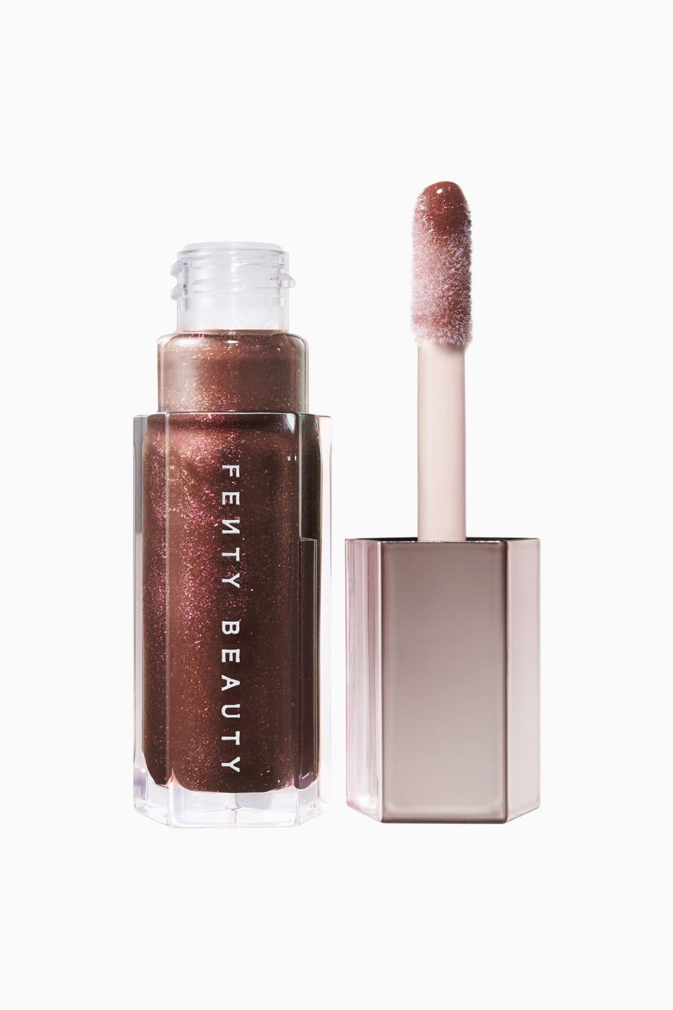 Näytä suurempi kuva: Avoin, kirkas kuusikulmainen Fenty Beauty -huulikiiltopullo seisoo pystyssä, ja sen sisältä näkyy hohtava syvä pronssi-ruskea sävy; tuotemerkki on painettu valkoisella pystysuoraan. Pullon vieressä lepää sen metallinhohtoinen ruusukullanvärinen kuusikulmainen korkki, jonka suuri, kiiltopäällysteinen doe-foot-applikaattorivarsi osoittaa ylöspäin.