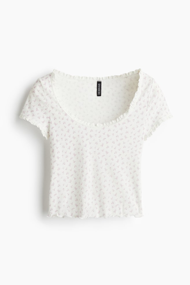 A short, fitted top in soft, white ribbed jersey is presented from the front, featuring a scattered pink floral pattern. It is designed with a round, lace-trimmed neckline, short sleeves with lace-trimmed cuffs, and an overlocked edge at the hem. - 1