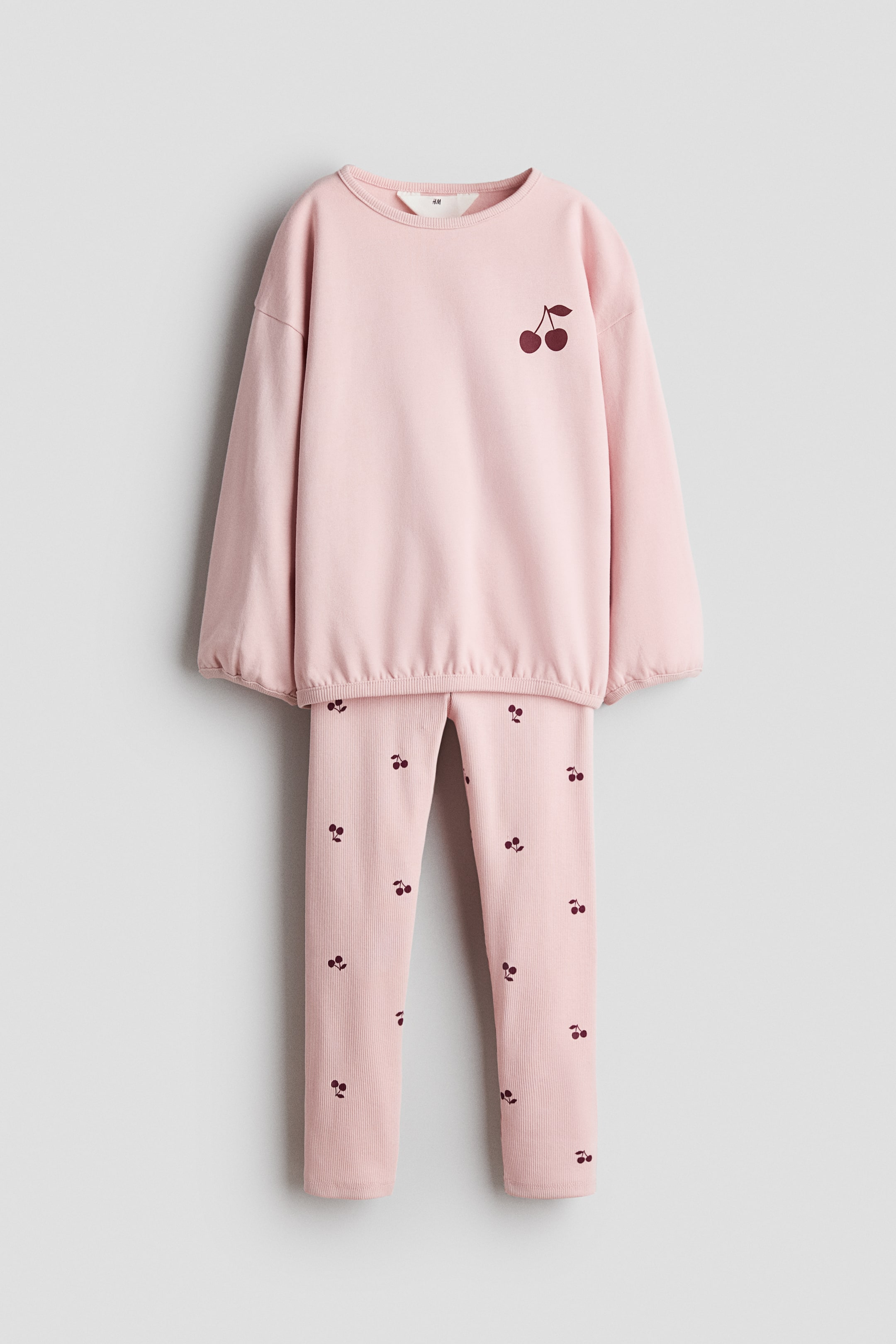 Ver imagen más grande: Un conjunto infantil de dos piezas en color rosa presenta una sudadera de manga larga con cuello redondo acanalado, puños acanalados, dobladillo acanalado y un gráfico de cereza en color rojo oscuro en el lado izquierdo del pecho. Los leggings a juego muestran una textura acanalada y un patrón de cerezas en color rojo oscuro en toda la prenda.