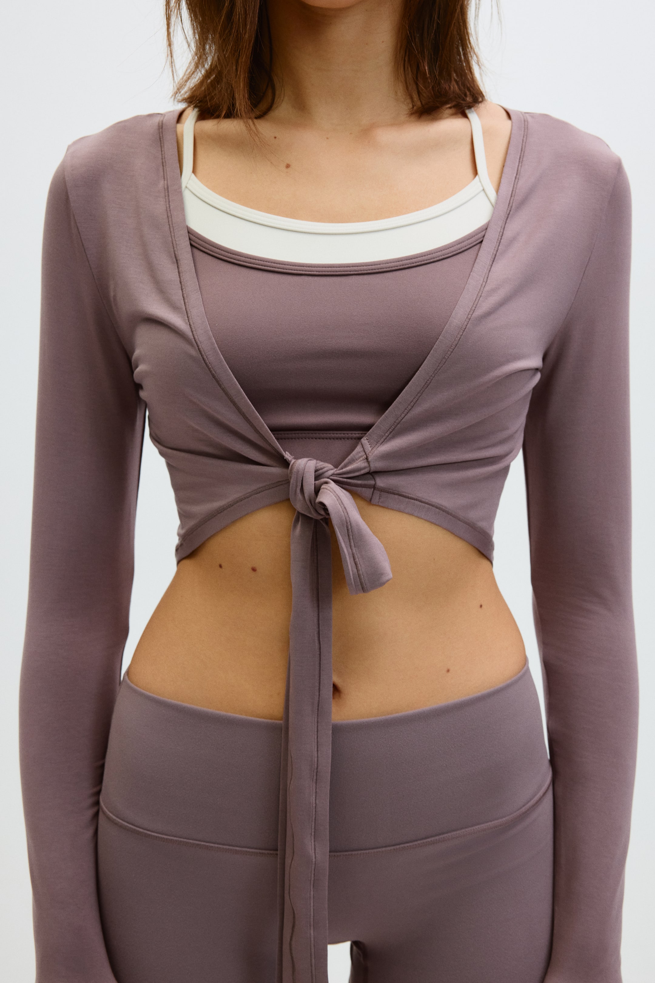 Ver imagen más grande: La modelo lleva puesto un top de yoga corto y ajustado en suave jersey de modal. Presenta un escote profundo en forma de V y mangas largas. El lazo al frente asegura el top en la cintura de la modelo, y el top está superpuesto sobre un top interior de color claro.