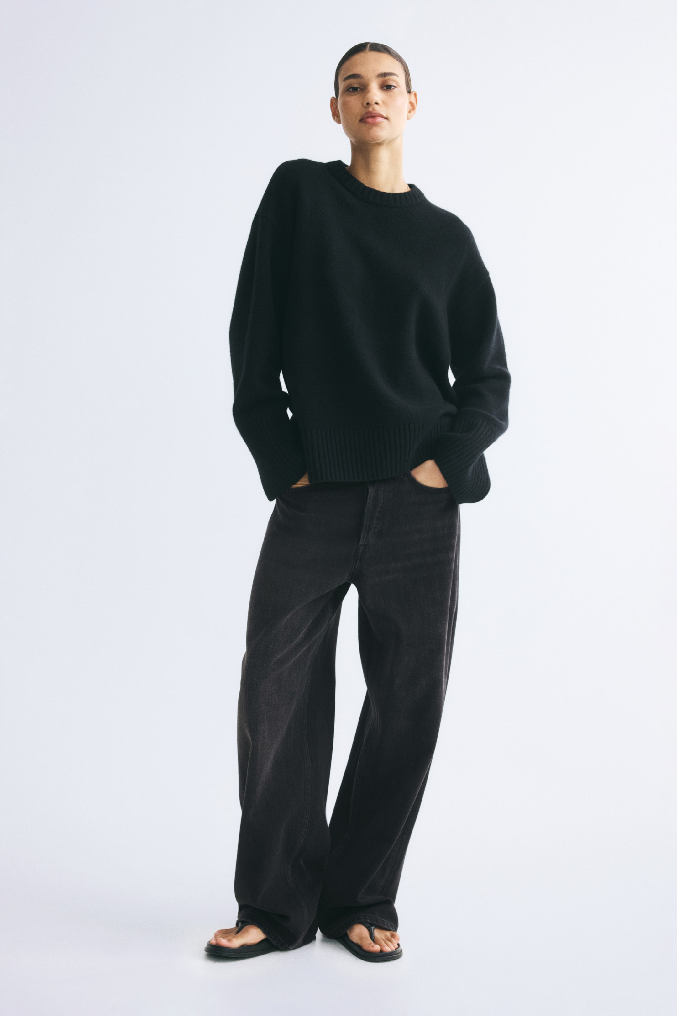 Agrandir l'image: Un jean en denim gris foncé, coupe ample et jambe droite, est porté par le mannequin, qui se tient debout les mains dans les poches. Le jean est associé à un pull noir à manches longues et col rond, ainsi qu’à des sandales noires.