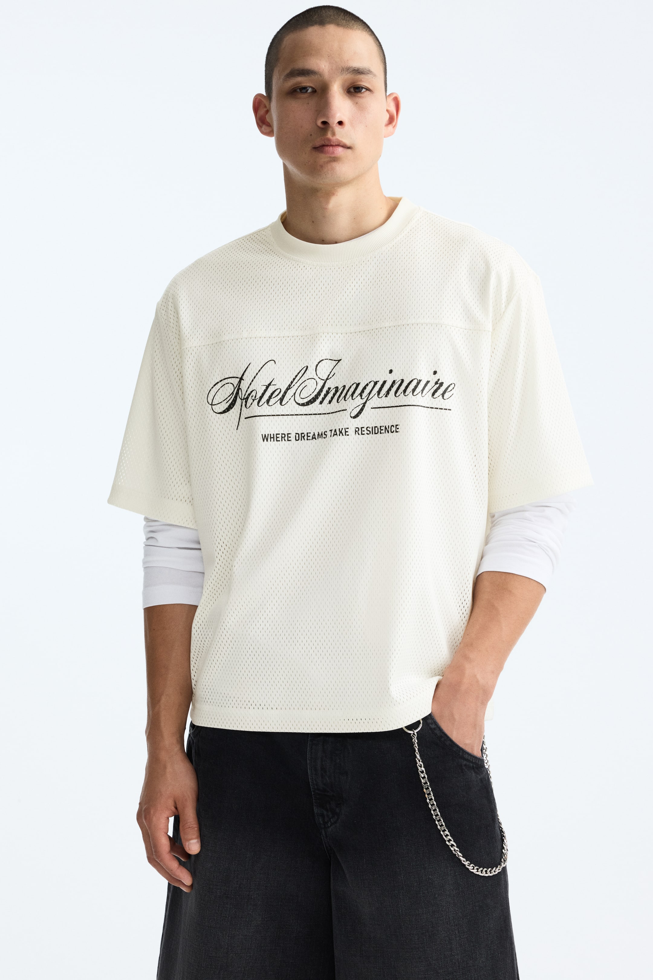 View larger image: The model is donning a cream T-shirt crafted from printed mesh, adorned with a black graphic design. It features a round neckline, dropped shoulders, a front yoke, and a straight-cut hem. The T-shirt exhibits an oversized fit, offering a baggy, extra-loose silhouette, and is stylishly layered over a white long-sleeve shirt.