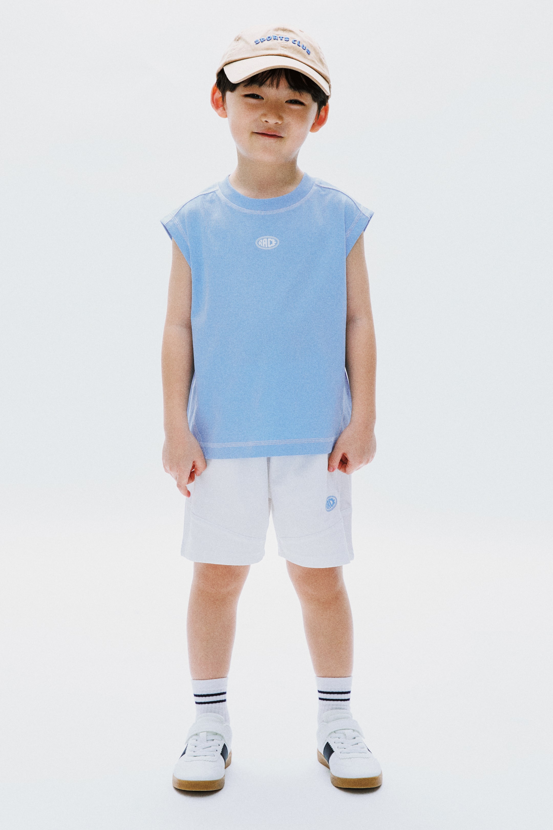 拡大画像を表示: 子供用アスレチックウェアのセットは、フロントに「SPORTS CLUB」の刺繍が施されたベージュのベースボールキャップ、クルーネックのライトブルーのノースリーブトップ、胸に白い楕円形の「R&D」ロゴが付いたデザインで構成されています。さらに、控えめな太腿のパネルと左脚にライトブルーの円形ロゴが入った白いショーツが含まれています。白いアンクルソックスには2本の黒いストライプがあり、フックアンドループファスナー付きの白いスニーカーには、ダークブルーのサイドストライプとライトブラウンのソールが施されています。すべてのアイテムは、正面からのビューで紹介されています。