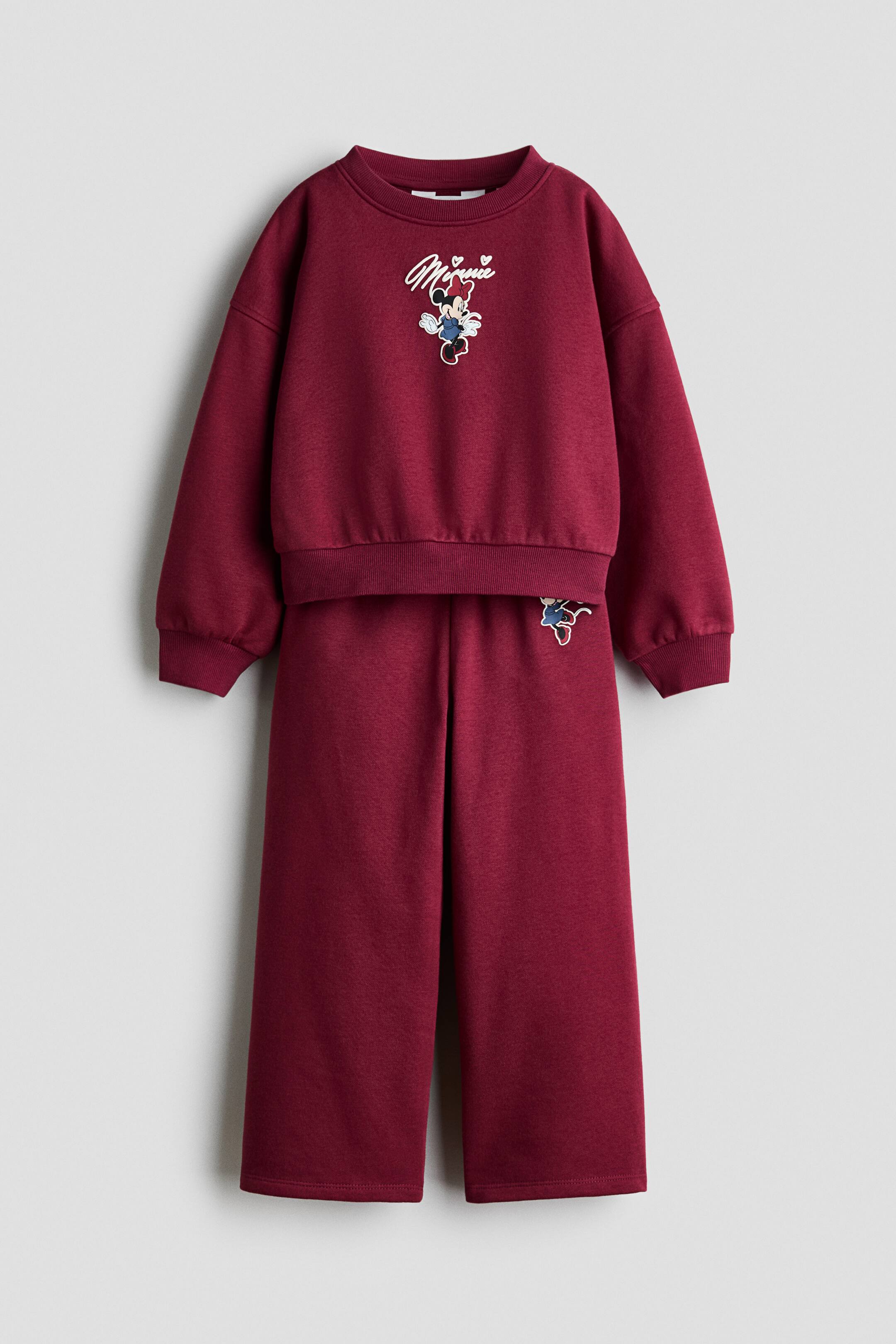 Prikaži veću sliku: Bordo dvodelni trenerka komplet je prikazan sprijeda i sastoji se od duksa sa okruglim izrezom i usklađenih pantalona širokih nogavica. Duks ističe centralni Minnie Mouse grafički motiv sa ispisom u kurzivnom vezu iznad, rebrastim manžetnama i rebrastim donjim rubom, dok pantalone prikazuju opuštenu siluetu sa manjim usklađenim motivom na gornjem levom delu butine.