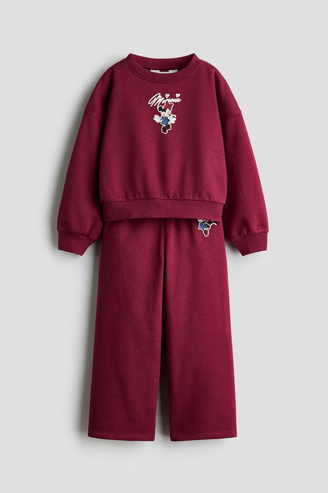 A burgundy two-piece tracksuit is shown from the front, consisting of a crew-neck sweatshirt and matching wide-leg trousers. The sweatshirt features a central Minnie Mouse graphic with cursive embroidered text above, ribbed cuffs, and a ribbed hem, while the trousers display a relaxed silhouette with a smaller matching graphic on the upper left thigh. - 1