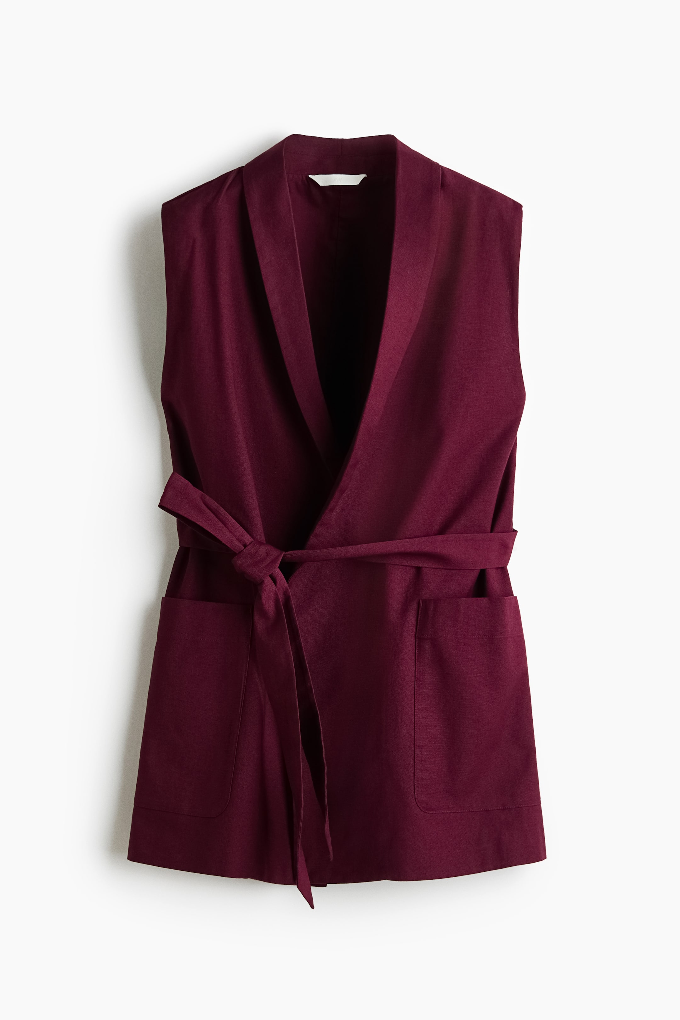 View larger image: A long, deep burgundy waistcoat is shown from the front, featuring a shawl collar and a wrapover front cinched at the waist with a tied belt. Two open front pockets are visible below the belt.