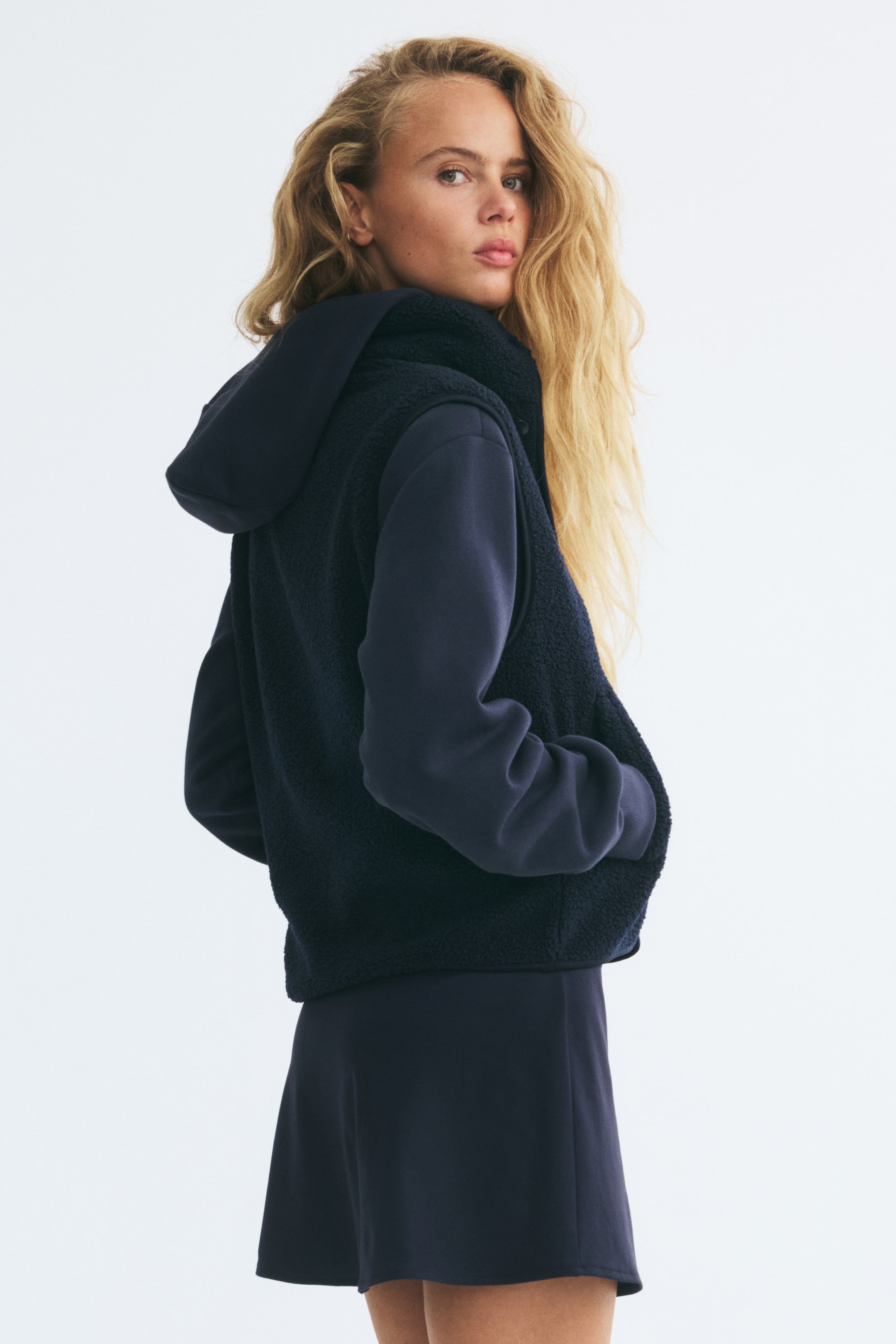 View larger image: A navy blue teddy fleece vest is being worn by the model over a matching long-sleeve hooded sweatshirt and skirt. The vest features a high funnel collar, jersey-bound edges, and diagonal front pockets in which the model is resting their hands.