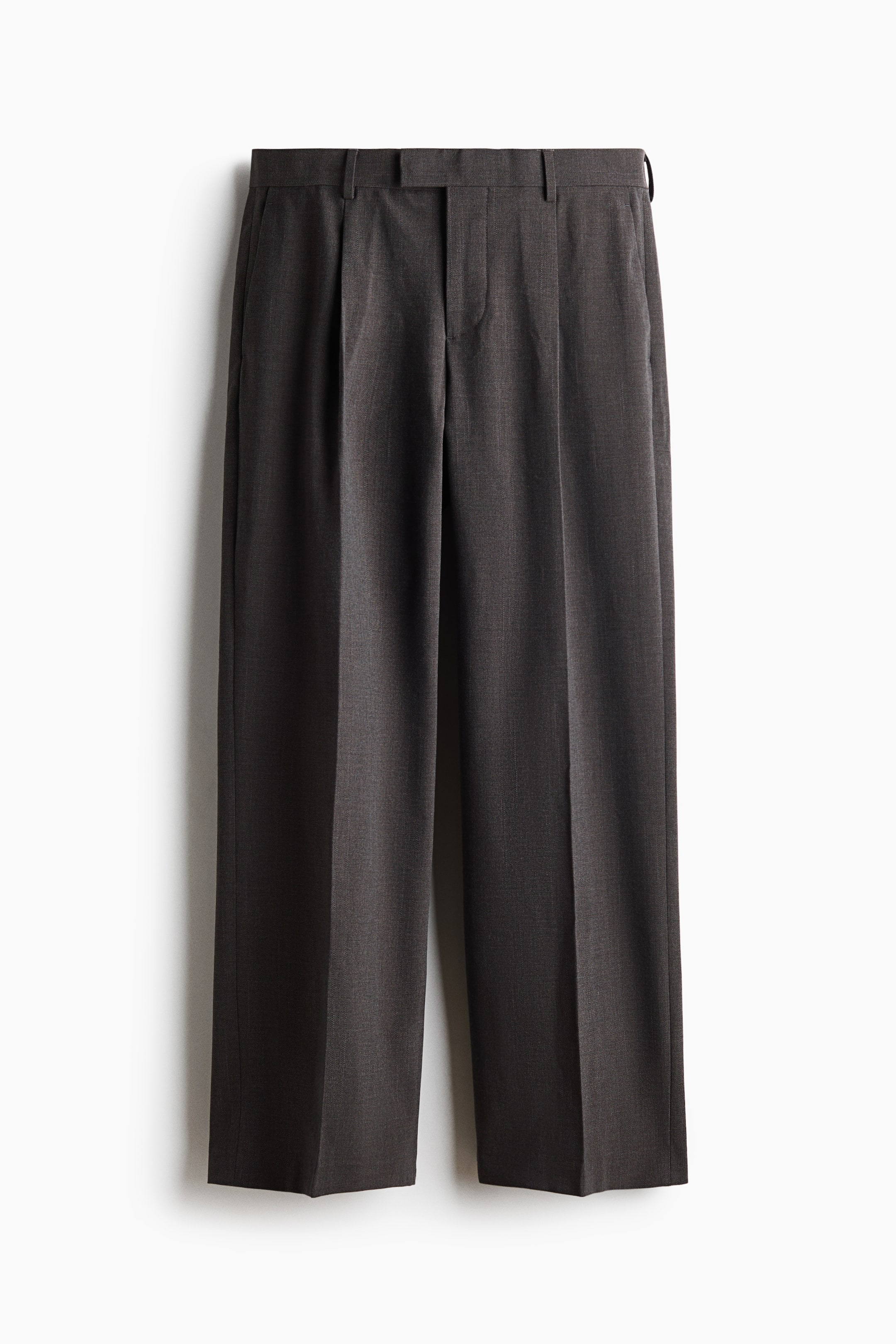 View larger image: Dark gray suit trousers are shown from the front, featuring an extended waistband with a zip fly closure. They include front pleats, side pockets, and straight legs with creases running down the front, contributing to a loose fit.