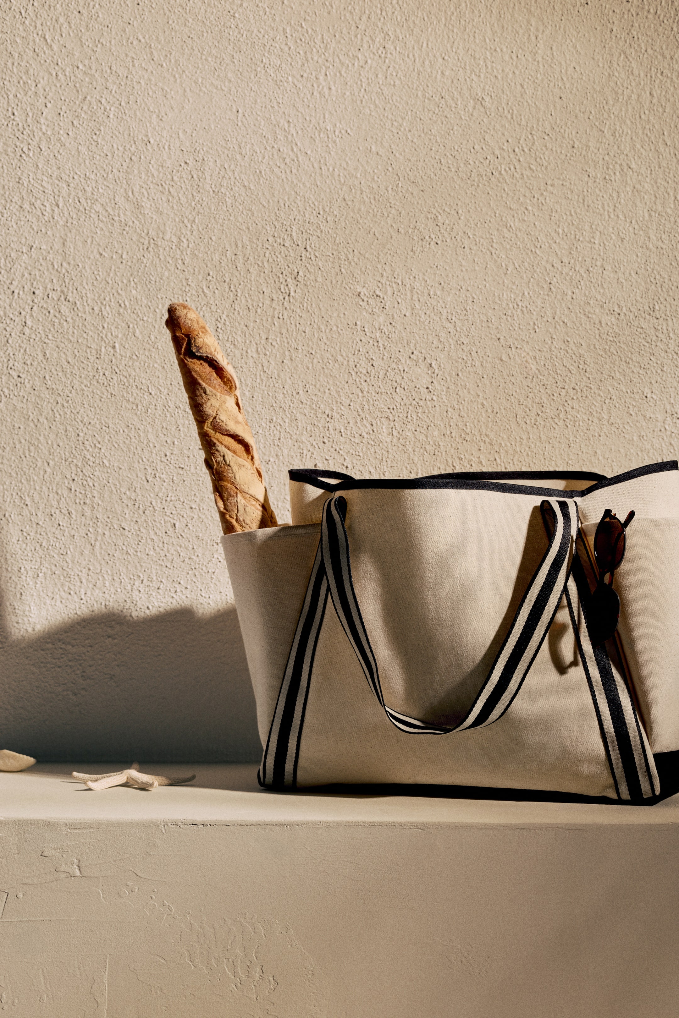 Ver imagen más grande: Una bolsa tote de lona crema está colocada, orientada ligeramente hacia la derecha, con correas de hombro a rayas negras y crema y un ribete negro a lo largo de la abertura superior. Un baguette está de pie dentro de la bolsa, y unas gafas de sol cuelgan del borde.