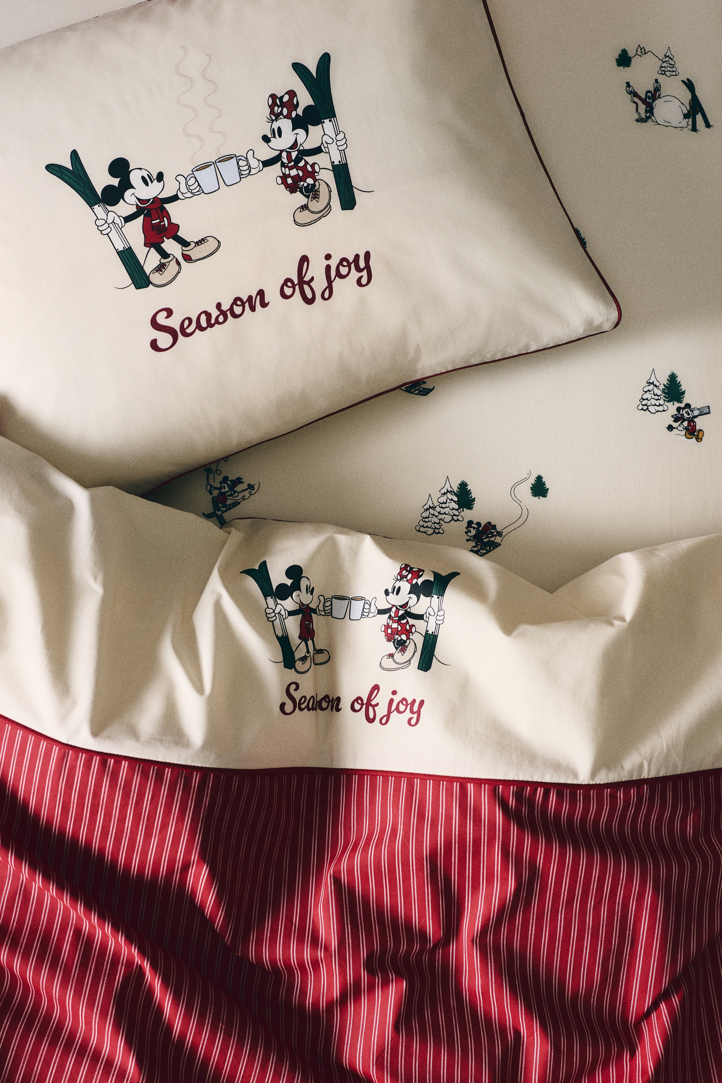 A cream-colored bed sheet is displaying a printed winter-themed pattern featuring Disney characters. The design shows Mickey Mouse skiing, Pluto wearing a red and white striped scarf on a sled, and sketch-style pine trees scattered across the off-white background.