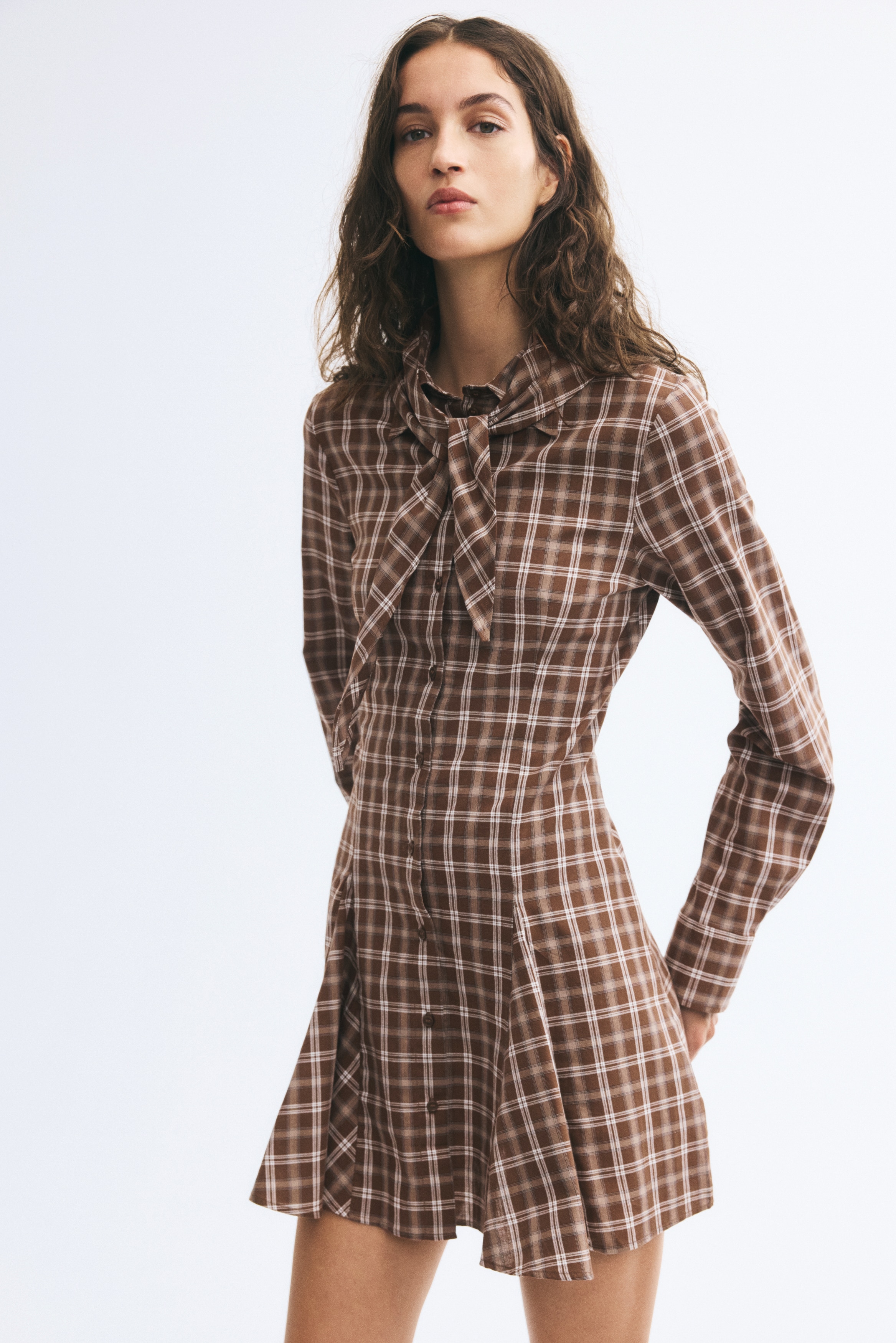 A brown plaid mini dress is shown from the front, featuring a button-up front, a tied bow collar, and long sleeves with wide cuffs. The dress is designed with a fitted bodice that flows into a flared, pleated skirt.
