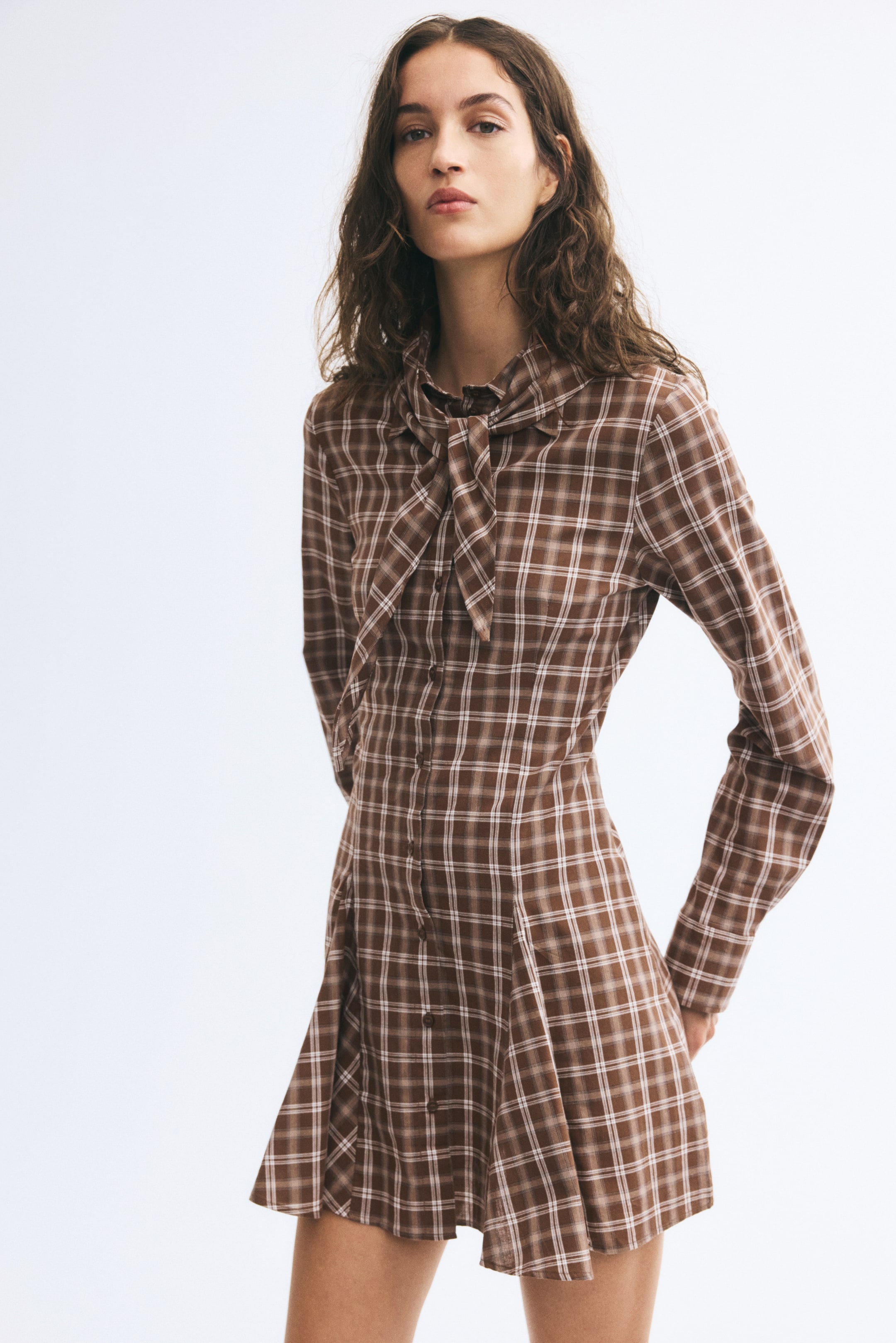 View larger image: The model is wearing a short brown-and-white plaid dress, featuring a button-down front, long sleeves, and a collar with a matching tied scarf. The dress has a fitted bodice with waist darts that flares out into a short skirt with godet pleats.