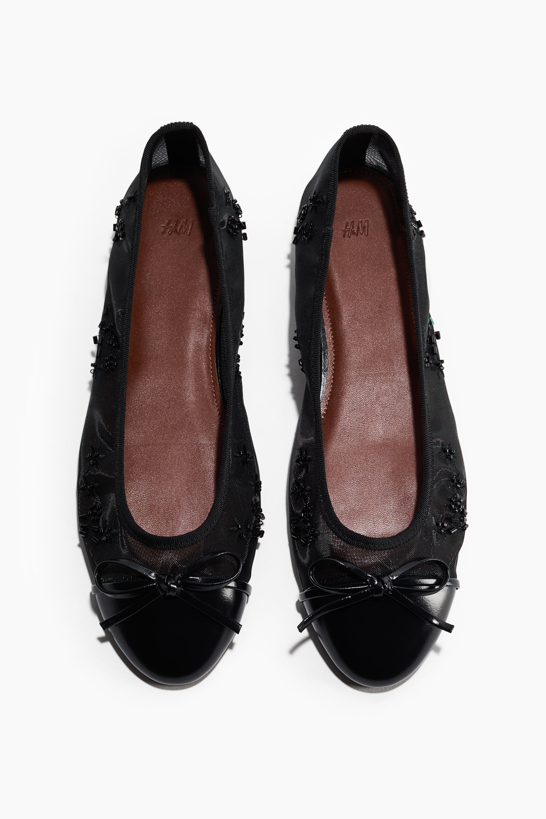 View larger image: A pair of black ballet flats is shown from a top-down, front-facing perspective, featuring semi-sheer mesh uppers embellished with small black beads along the sides. The shoes showcase glossy patent leather toe caps topped with delicate bows and contrasting brown insoles.