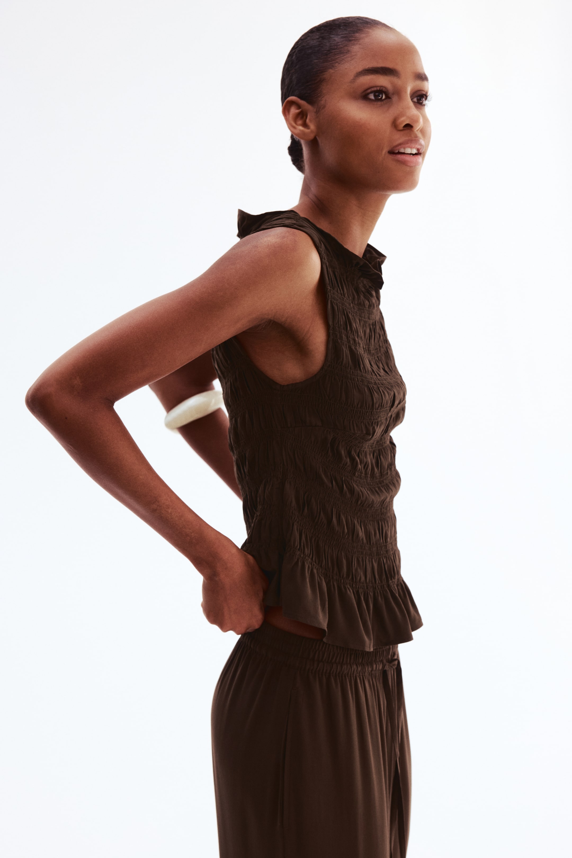 View larger image: A fitted, sleeveless top in dark brown smocked viscose weave, featuring a frill-trimmed boat neckline and a frill trim at the hem, is worn by the model and styled with matching pants. The model is adjusting the top at the waist.