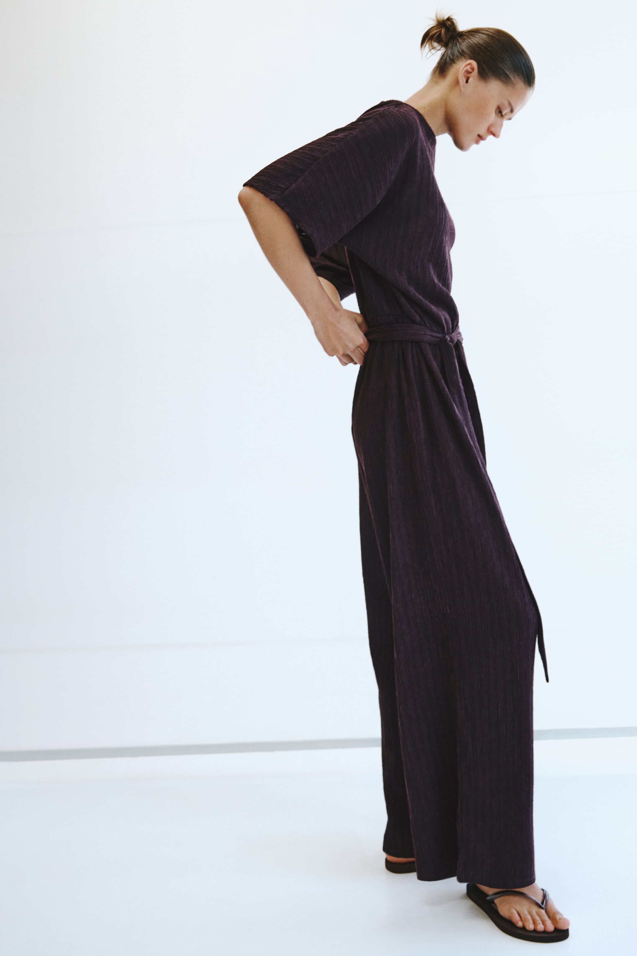 Größeres Bild ansehen: Ein dunkelvioletter Jumpsuit mit dezenten vertikalen Falten wird aus einem seitlichen Rückwinkel gezeigt. Er verfügt über kurze Dolman-Ärmel, einen Gürtel aus demselben Stoff und weit geschnittene Hosenbeine.