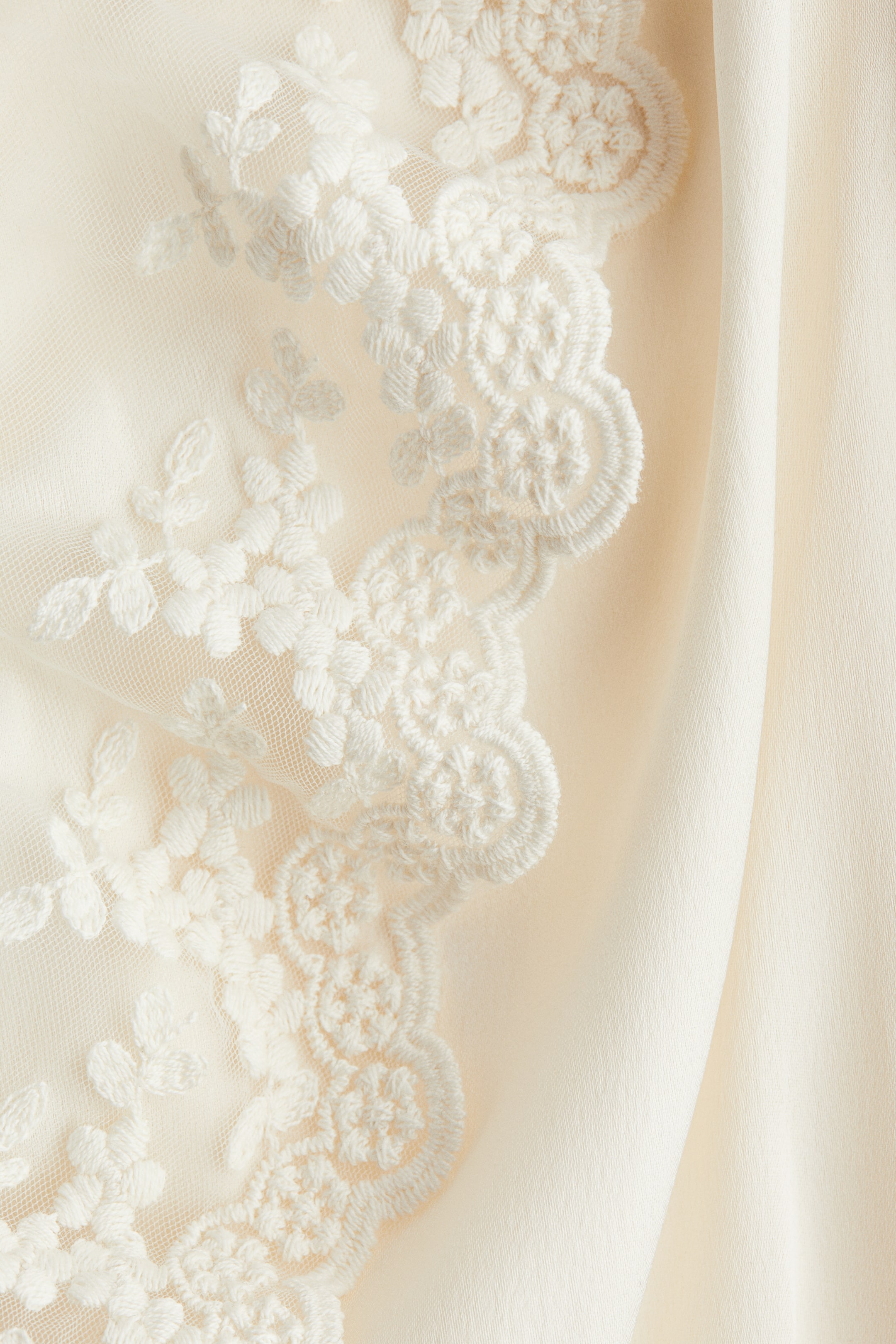 View larger image: A cream-colored sheer fabric is featuring intricate white floral and swirling embroidery along a scalloped edge, lying adjacent to a smooth, solid cream-colored fabric.
