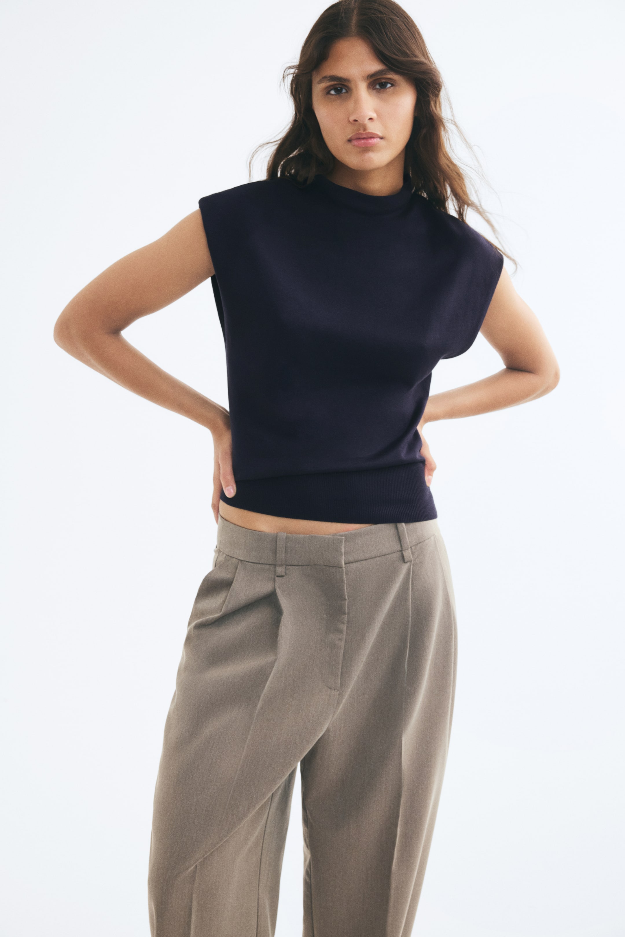 View larger image: A dark navy sleeveless knit vest with a high neck and a ribbed hem is worn by the model. The vest is paired with pleated taupe trousers.