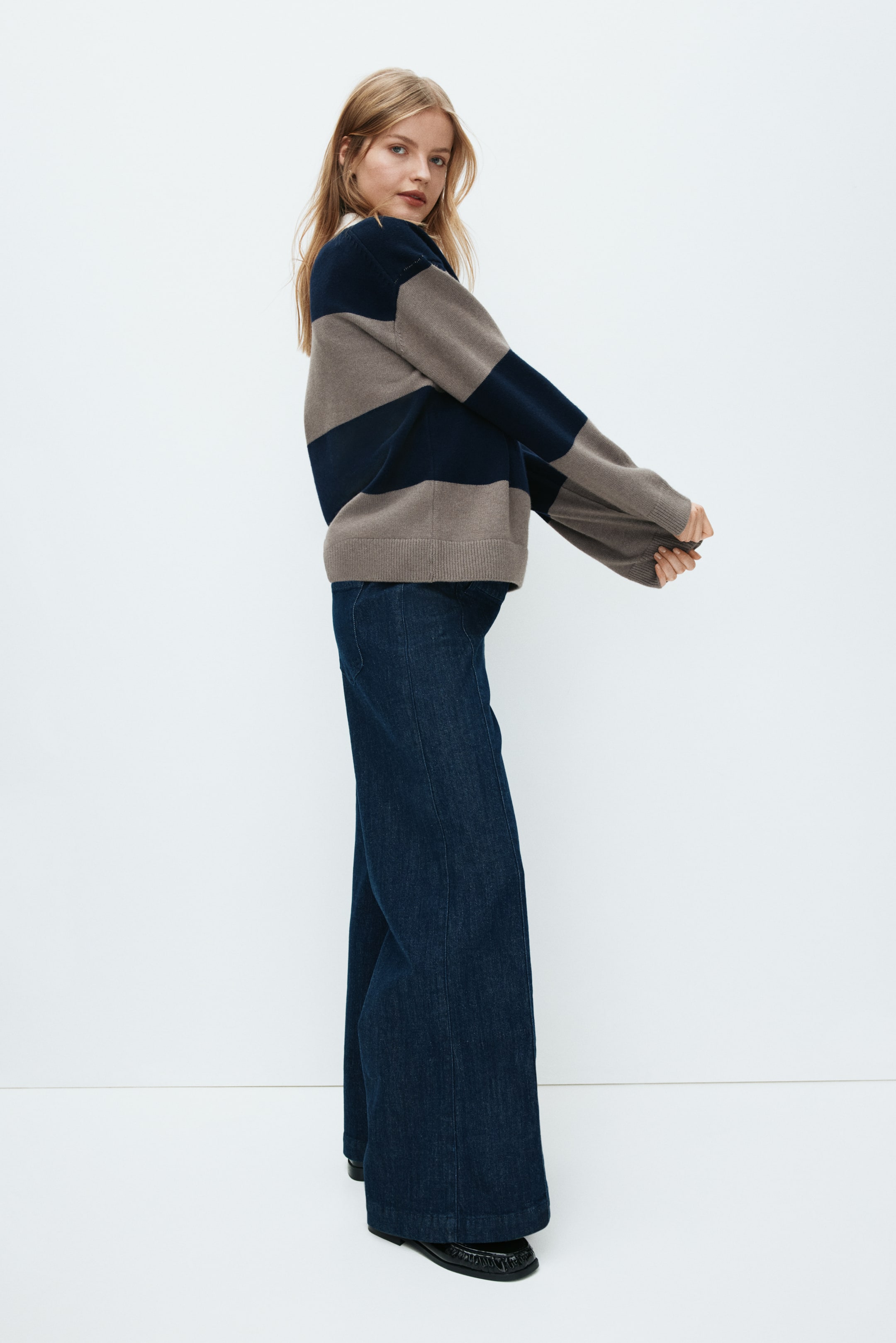 View larger image: A relaxed-fit jumper features wide horizontal stripes in dark navy and light brown, with a white collar visible beneath its crew neck. The long sleeves extend past the wrists, finished with ribbed cuffs, and it is styled with dark-wash, wide-leg jeans, shown from a side-front angle.