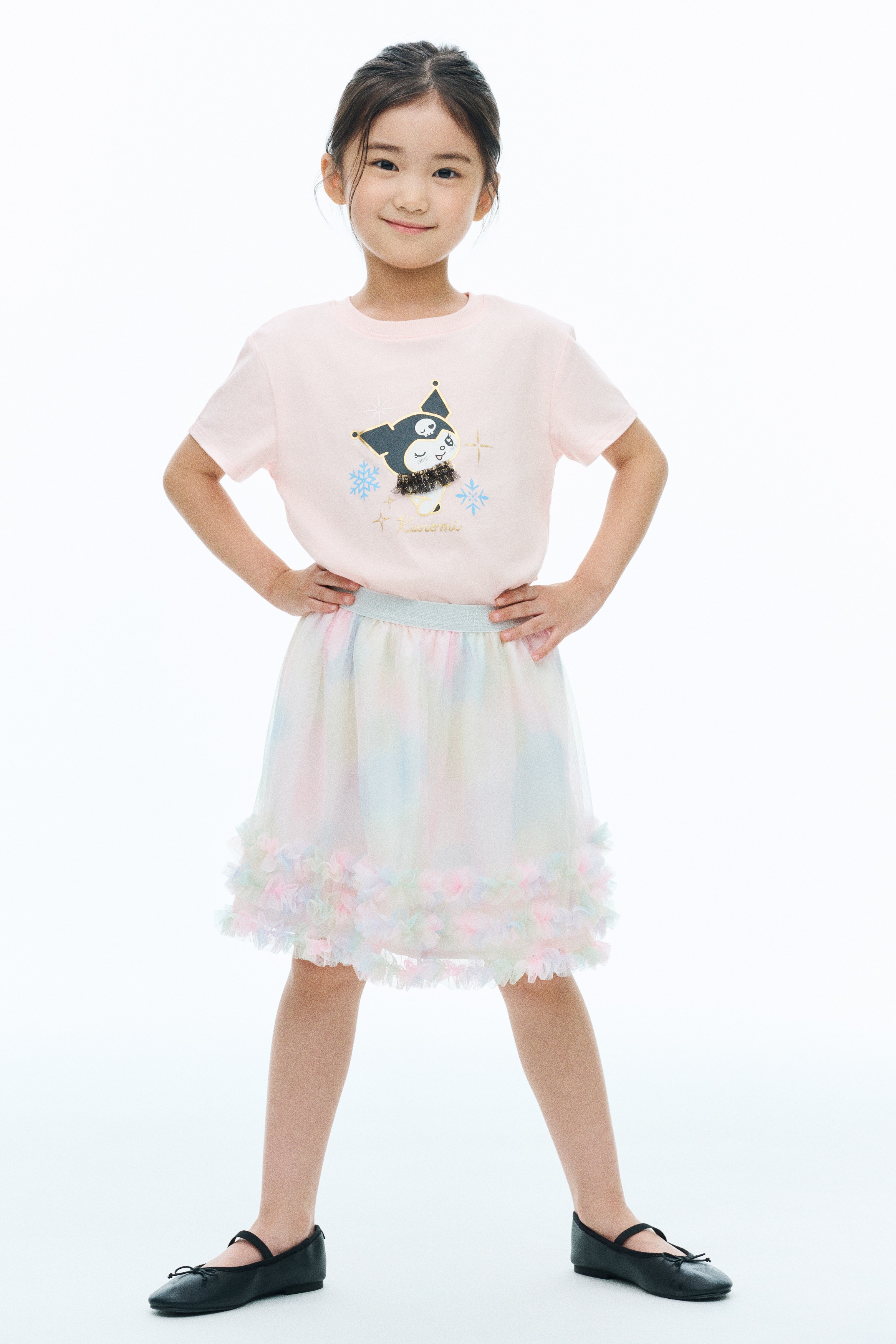 A light pink, short-sleeved T-shirt is shown from the front, featuring a ribbed crew neck and a central graphic design. The graphic features the character Kuromi wearing a black jester hat with gold foil accents and a black tulle collar, surrounded by blue snowflakes, gold sparkles, and the name âKuromiâ written in gold script below.