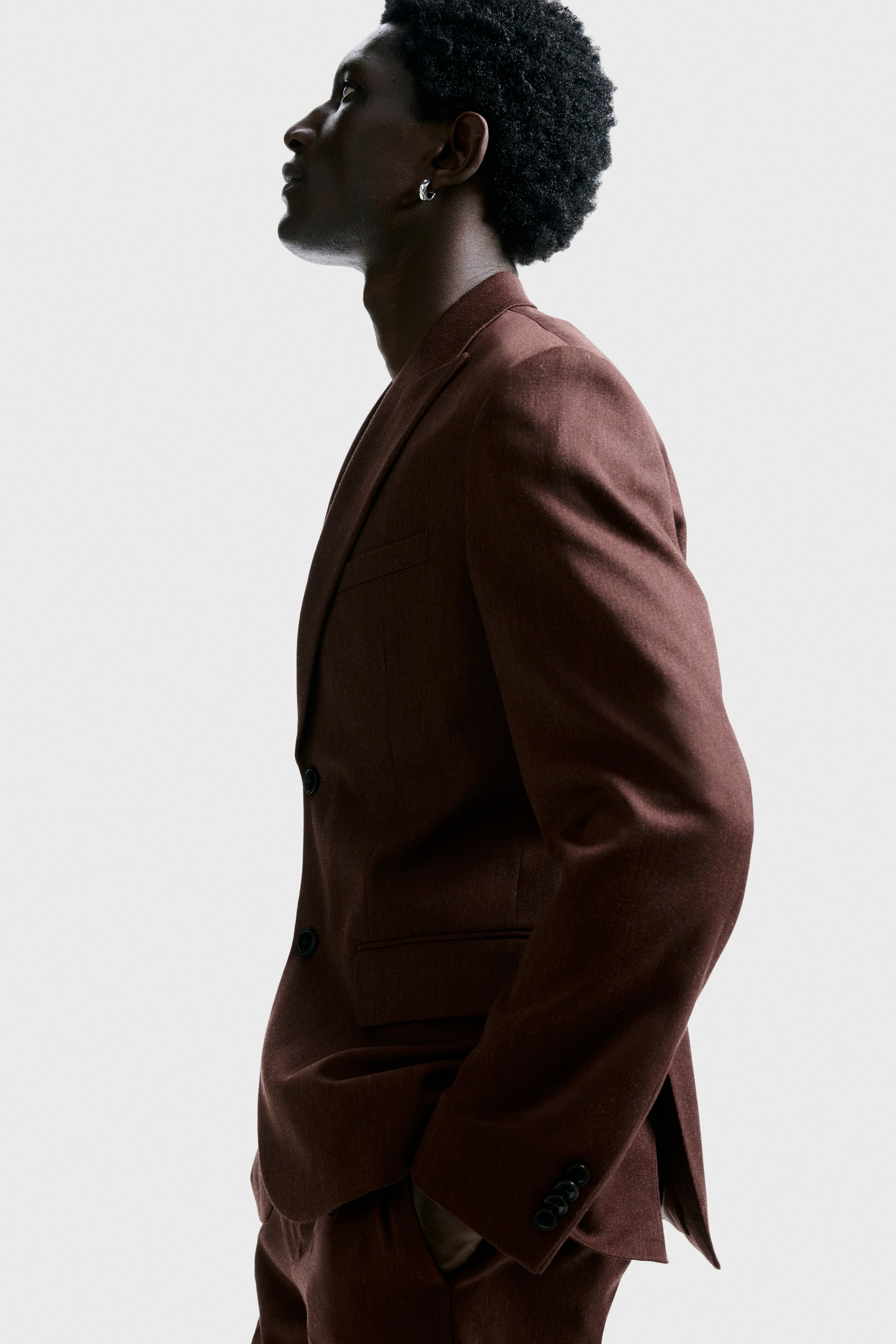 拡大画像を表示: 濃い赤褐色のスーツが側面から示されています。ジャケットは、ブラッシュされた質感、ノッチラペル、前面に2つのボタン、胸ポケット、フラップポケット、そして4つの袖ボタンを備えており、同じデザインのトラウザーも見えます。