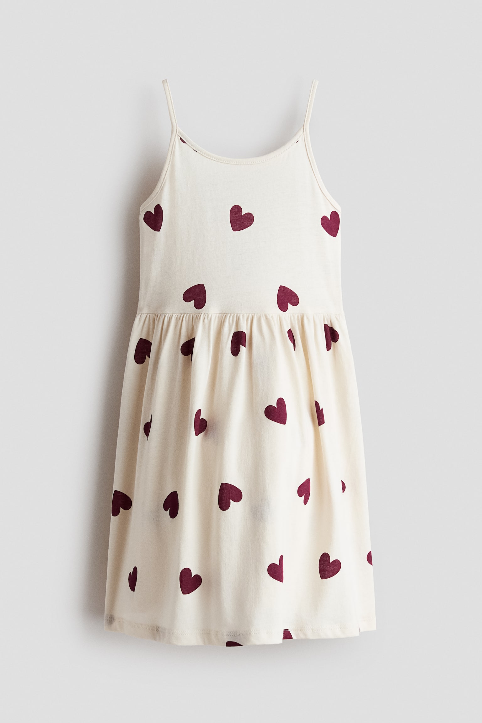 This front-facing cream-coloured camisole dress features an all-over pattern of dark red hearts. It showcases thin spaghetti straps, a scoop neckline, and a gathered waistline that flows into a flared skirt.