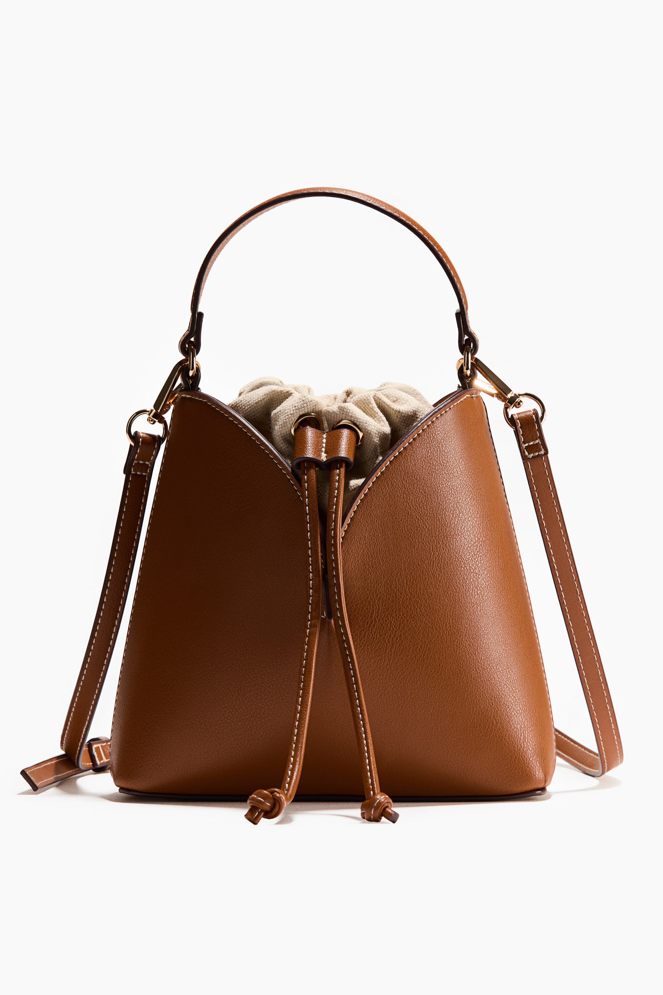 View larger image: A brown bucket-style handbag is presenting front facing, featuring a flared body with white contrast stitching and gold-toned hardware. A light beige fabric drawstring closure is cinching at the top, secured by brown leather drawstrings, and a curved top handle and detachable shoulder strap are attaching.