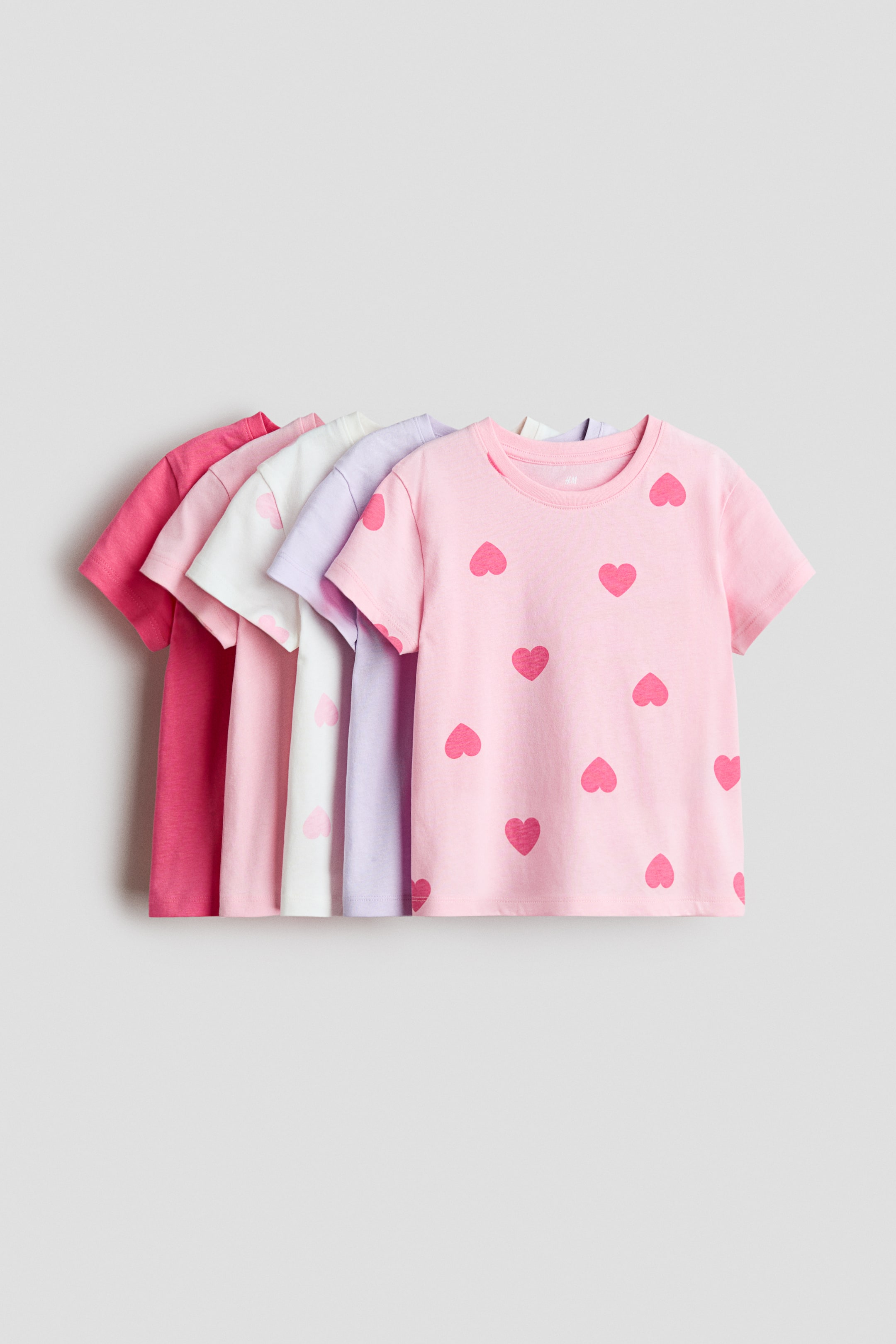 View larger image: Five jersey T-shirts are arranged in a fan shape, each boasting a round, ribbed neckline and subtly dropped shoulders. The foremost light pink T-shirt showcases a repeating pattern of darker pink hearts, while the shirts behind it appear in solid shades of pink, white, and lilac.