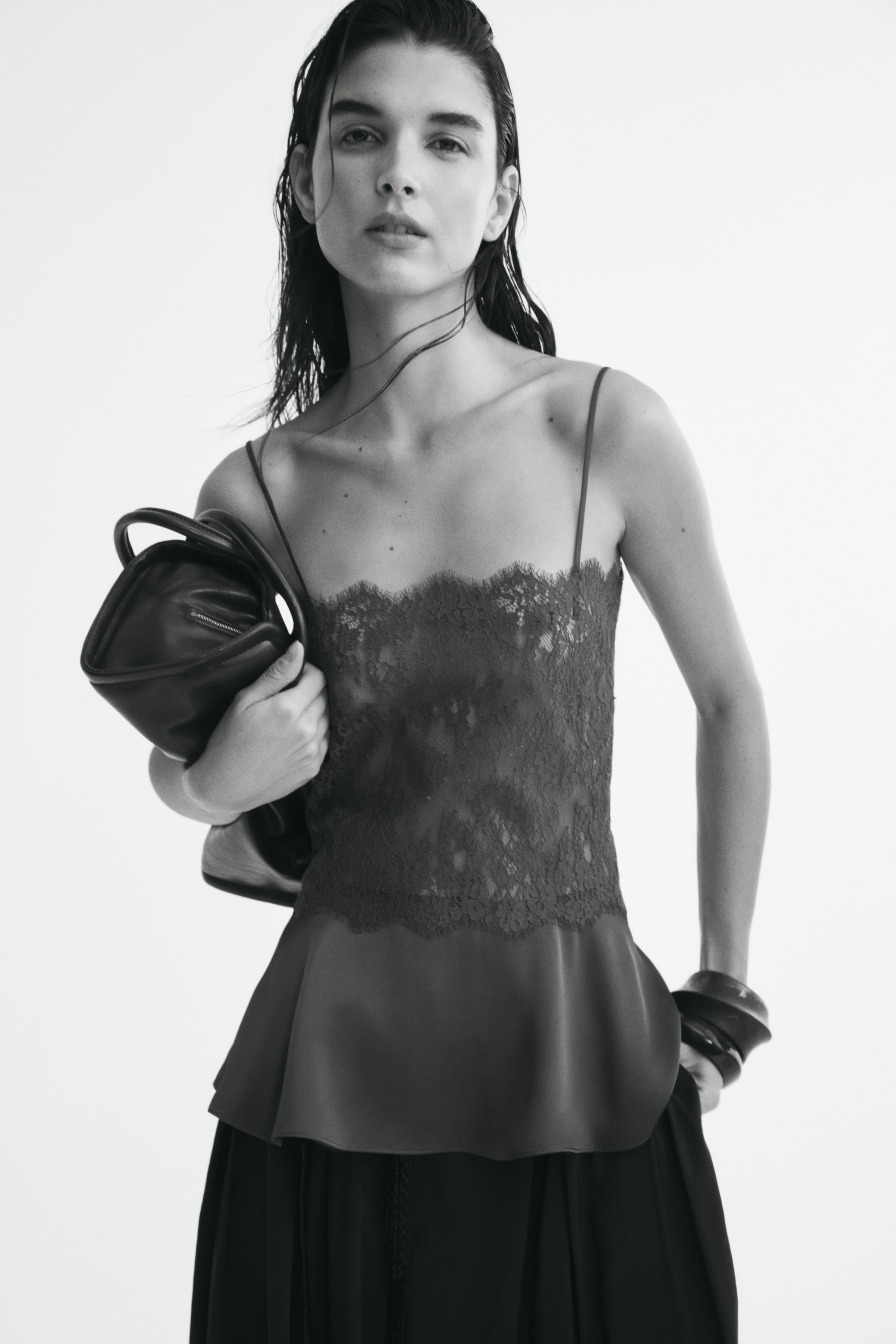 View larger image: A fitted lace camisole featuring narrow shoulder straps, a straight scalloped neckline, and a flared peplum hem is being worn by the model. The model is holding a dark leather bag under one arm and wearing dark trousers.