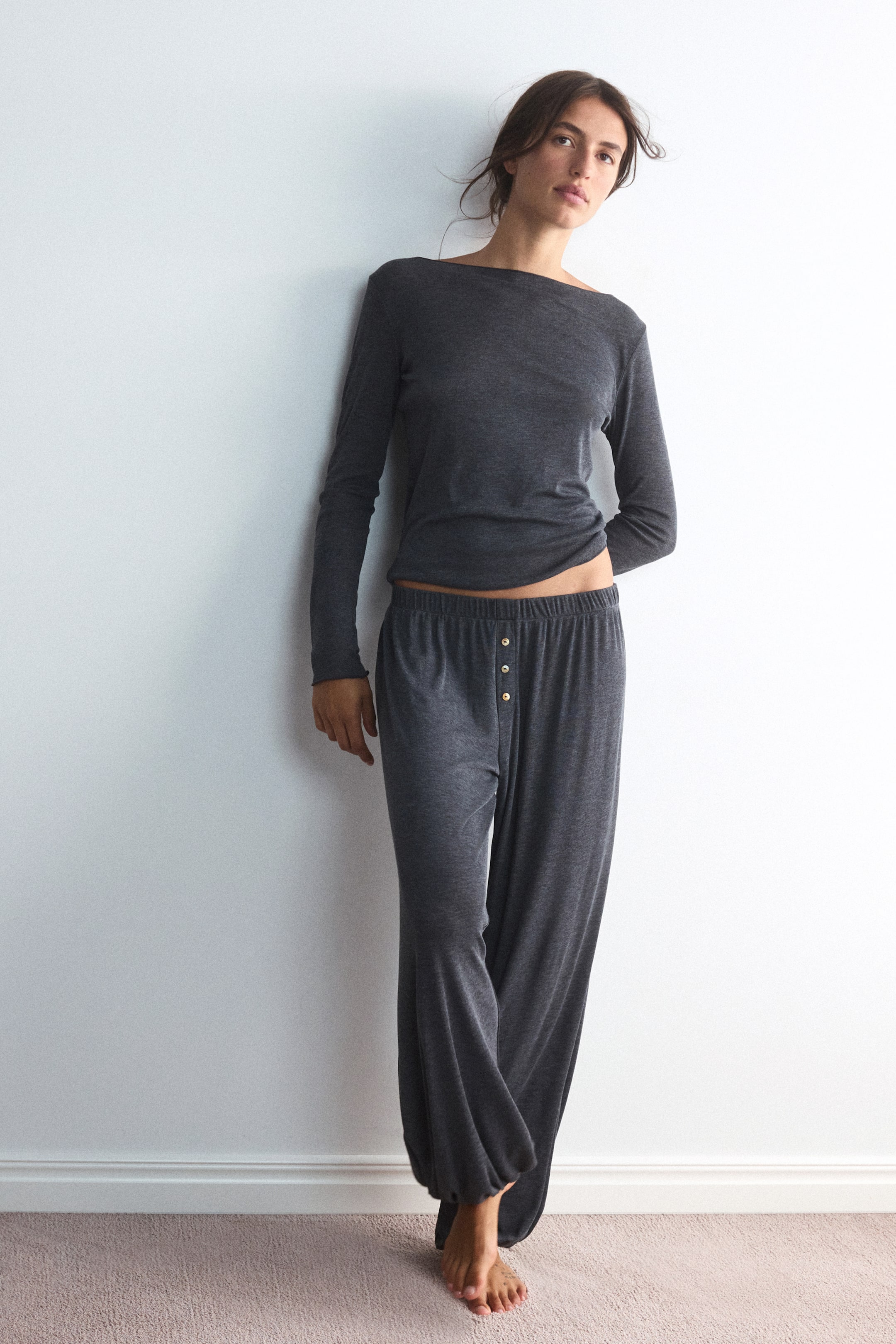 View larger image: The model is wearing a dark grey jersey pyjama set, featuring a long-sleeved top with a boat neckline and matching loose-fit pants. The pants come with an elasticated waistband, a mock fly with three small decorative buttons, and balloon-style legs with elasticated hems.