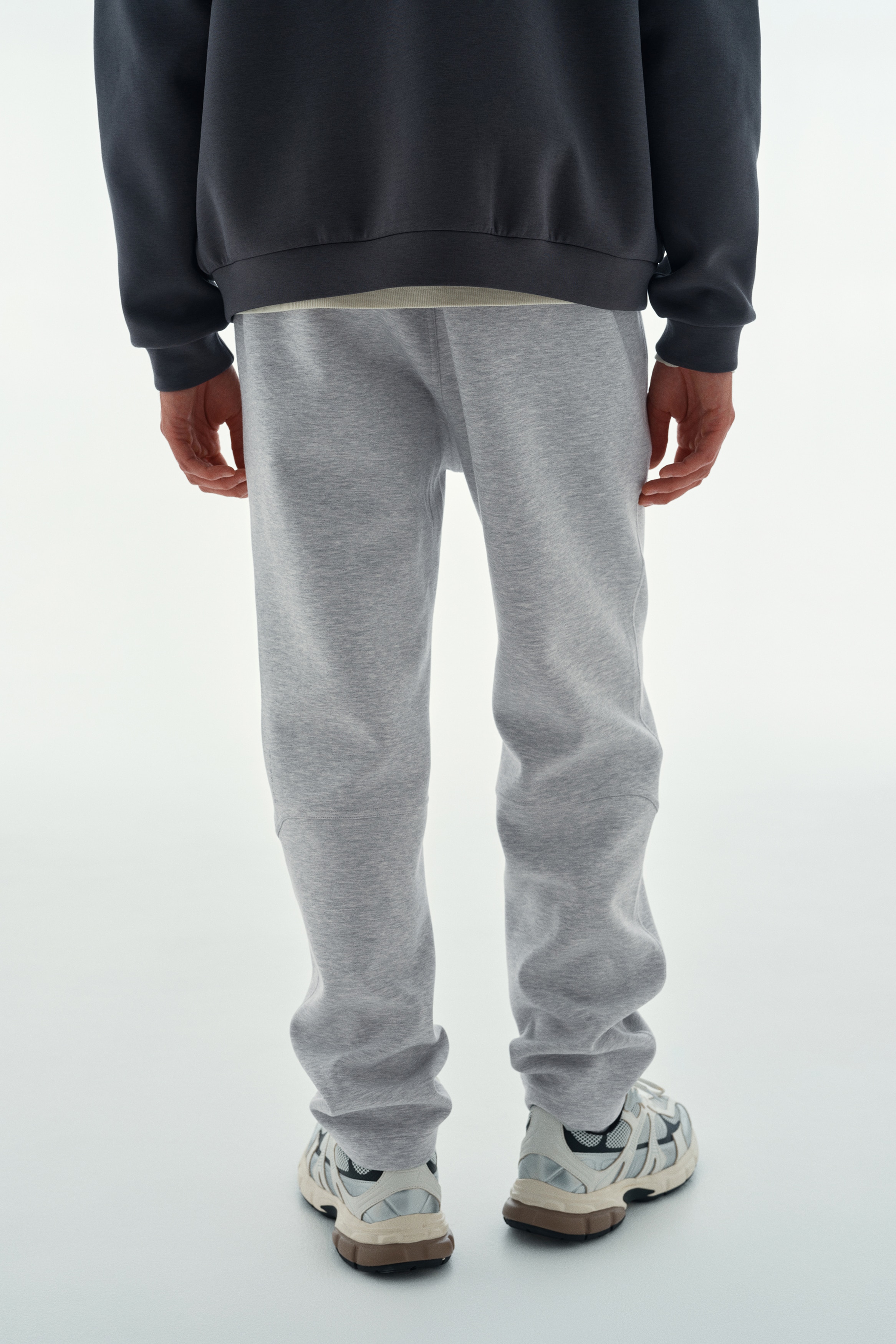 Hellgraue Jogginghose von vorne, mit elastischem Bund und schrägen Seitentaschen. Vertikale Nähte verlaufen entlang der äußeren Beine und enden in schlichten Säumen.