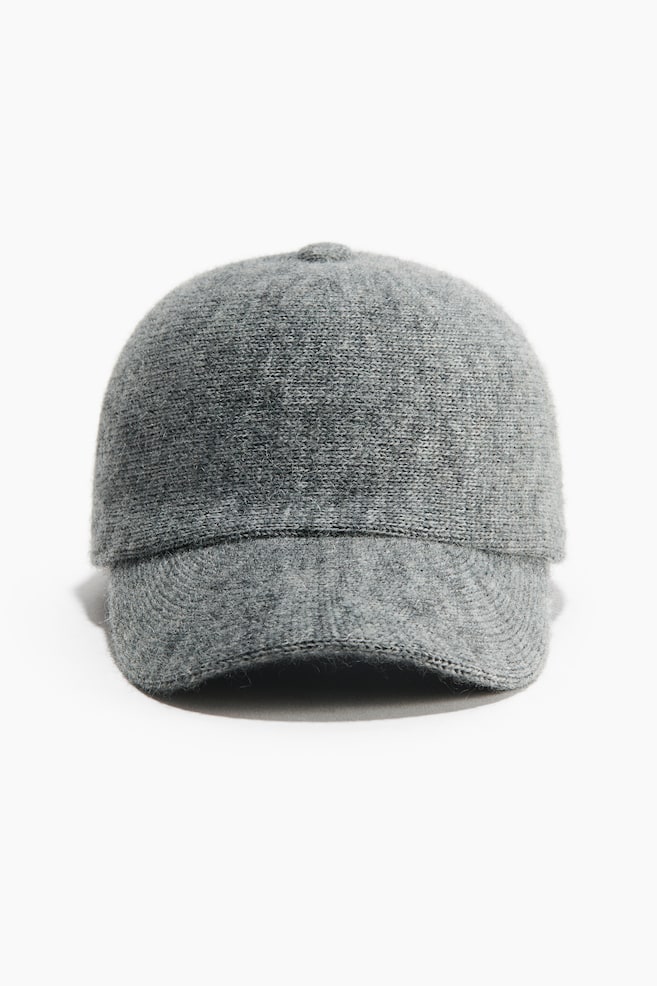 Une casquette de baseball en maille grise est présentée de face, mettant en valeur sa texture tricotée, sa calotte structurée et sa visière courbée. Un bouton assorti en maille est placé au centre supérieur de la casquette, soulignant son design doux et harmonieux. - 1