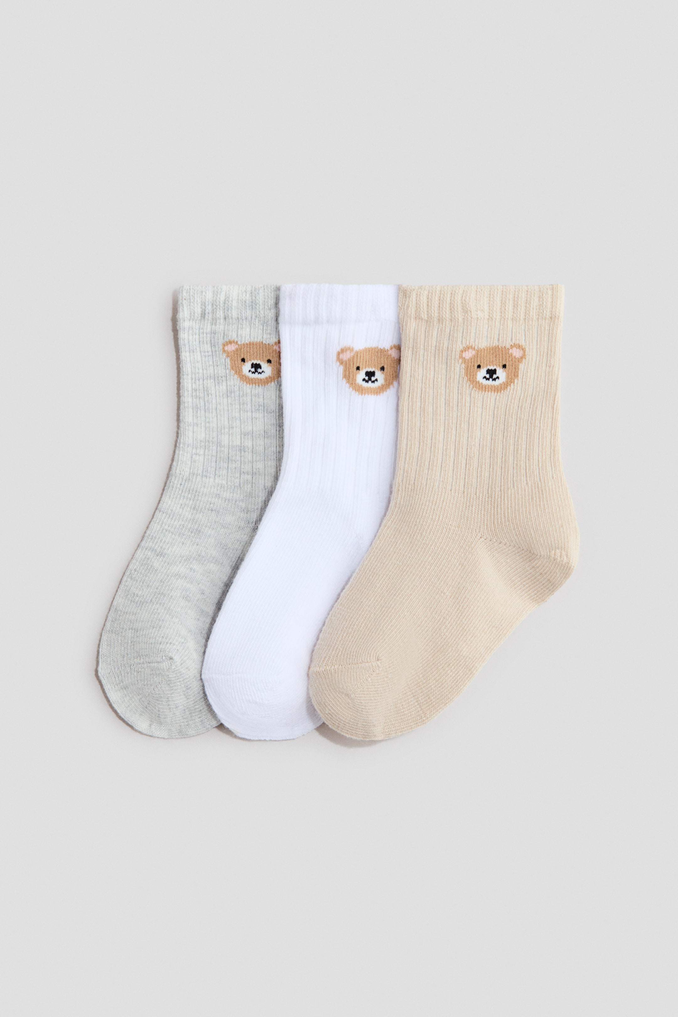 Xem ảnh lớn hơn: Ba chiếc tất dệt kim mịn trong hỗn hợp cotton mềm mại được trưng bày cạnh nhau, hướng về phía trước. Những chiếc tất có màu xám nhạt, trắng và be nhạt, mỗi chiếc đều có phần trên gân và một khuôn mặt gấu bông thêu.