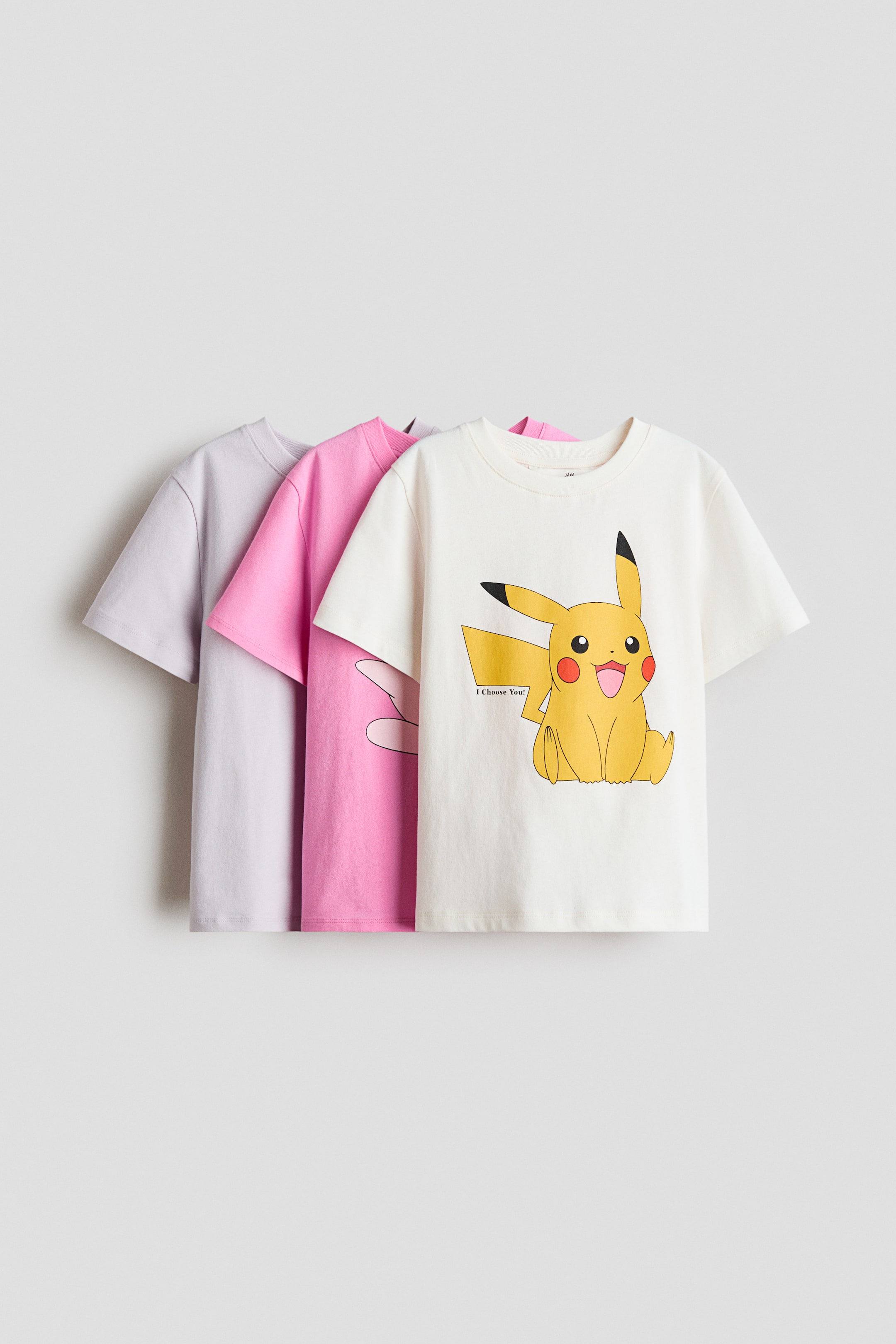 Voir une image plus grande: Un ensemble de trois hauts à manches courtes en jersey de coton doux est présenté en vue de face dans une disposition en couches. Le haut avant, de couleur blanc cassé, arbore un imprimé coloré d'un personnage de dessin animé jaune avec des oreilles à pointes noires et le texte « Je te choisis ! ». Un haut rose, partiellement visible derrière, présente le contour léger d'une oreille de personnage, avec un haut violet clair uni en arrière-plan.