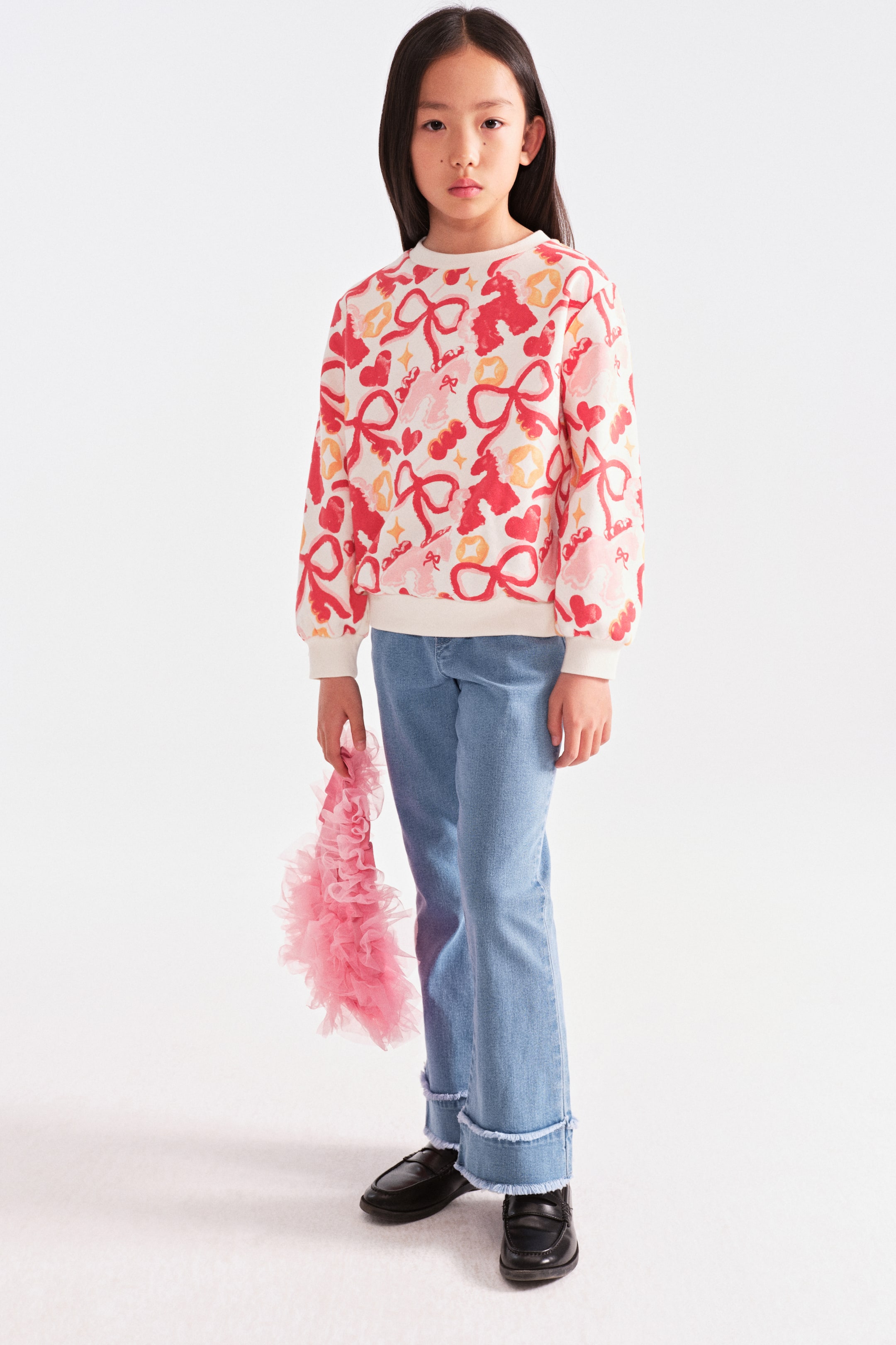 拡大画像を表示: 子どもがクリーム色の長袖クルーネックスウェットを着用しており、全体に赤いリボン、ピンクのハート、黄色のモチーフの総柄プリントが施されています。袖口と裾はリブ仕様で、ライトブルーのストレートジーンズ（裾はフリンジ加工の折り返し）と合わせています。